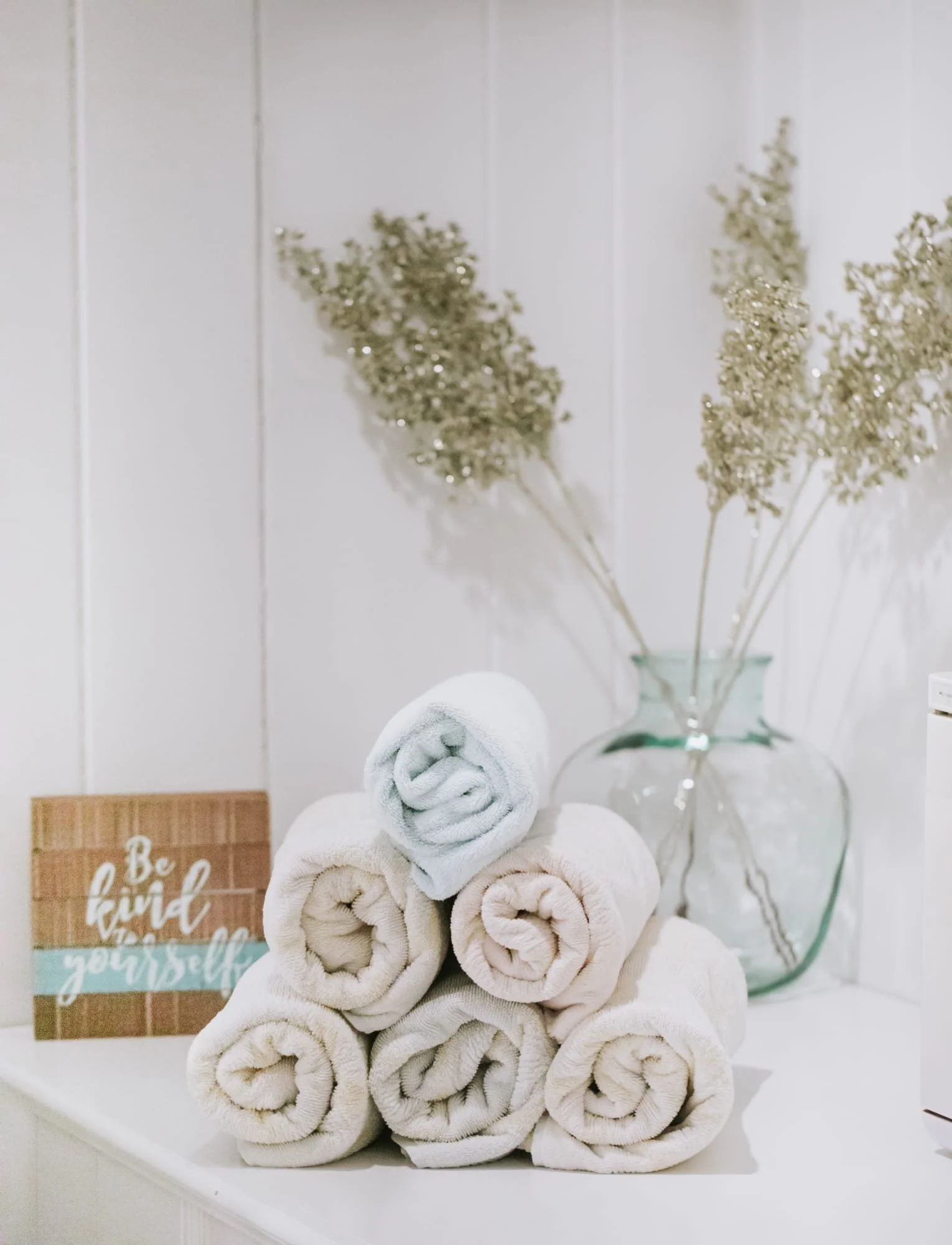 Stacked white and beige rolled towels on a white surface with a small wooden sign that says 'Be kind to yourself,' a clear glass vase with dried flowers, and a white wall in the background.