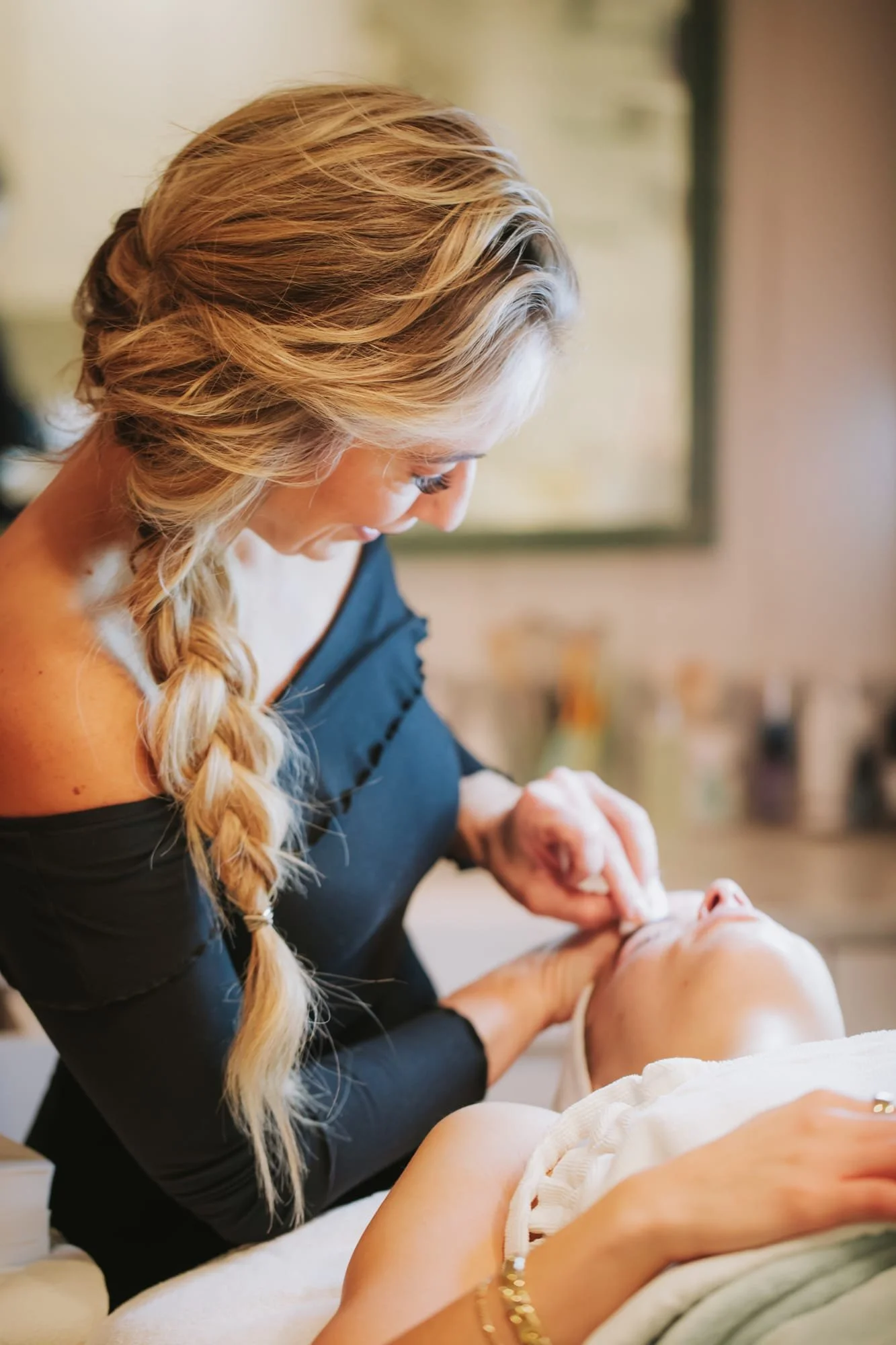 A woman with blonde hair in a braid, wearing a black off-the-shoulder top, is gently applying makeup or skincare to a woman lying down.
