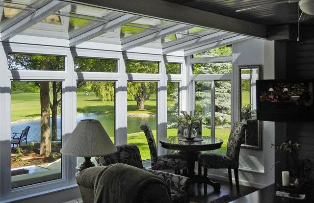 Interior view of a sunroom with large glass windows showing a view of a pond and trees outside, furnished with a round dining table, four chairs, a lamp, and decorative plants.