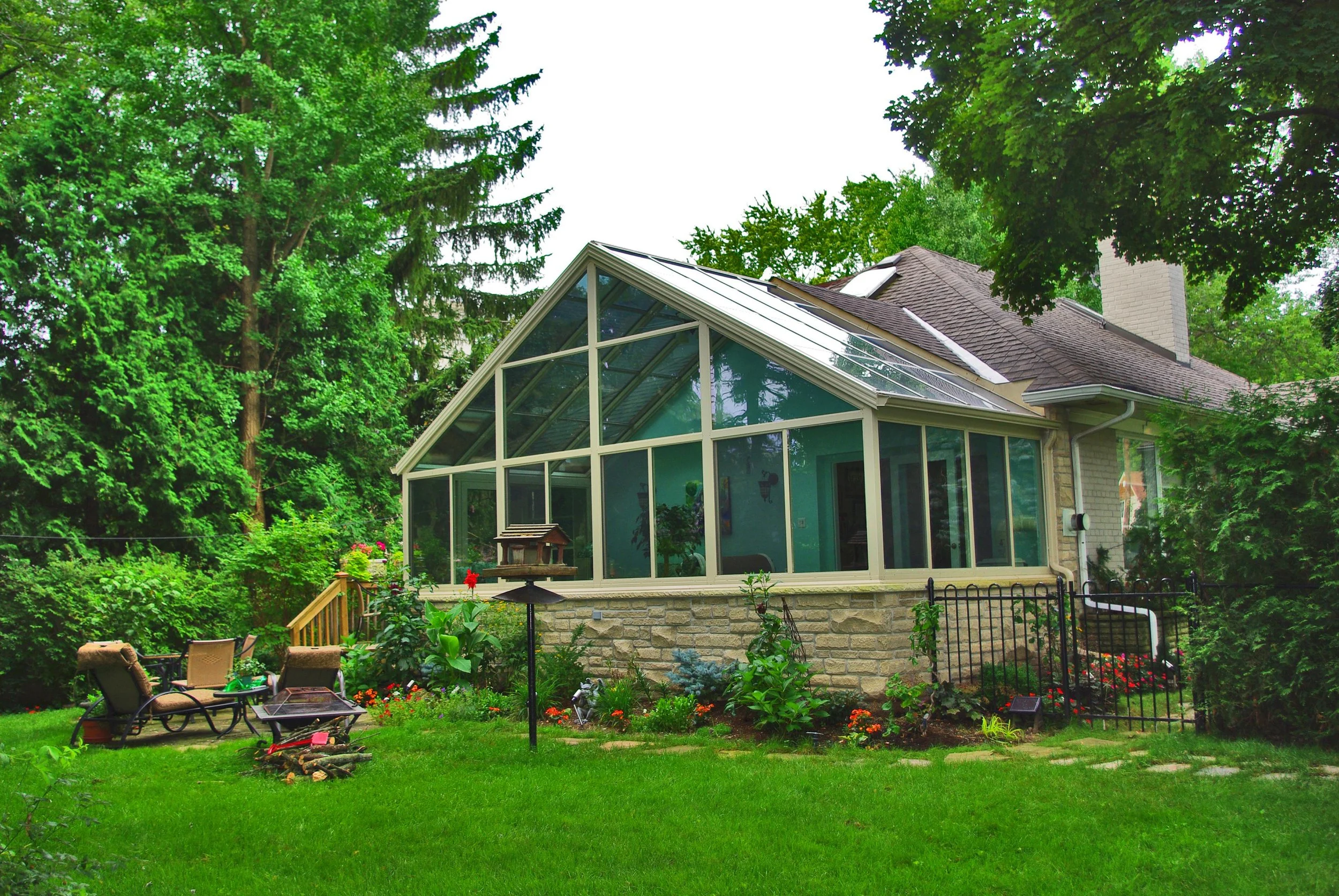 A backyard with a raised garden bed, outdoor chairs, and a fire pit, featuring a house with a glass sunroom and solar panels on the roof, surrounded by tall trees and lush green grass.