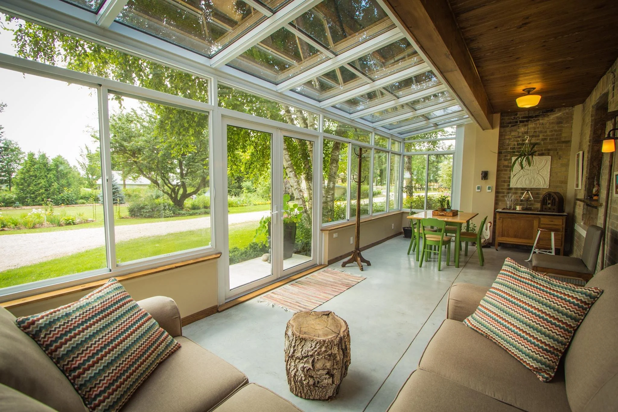 Sunroom with large glass windows and ceiling, green trees outside, beige sectional sofas with patterned pillows, a wooden coffee table, and a small dining table with green chairs.
