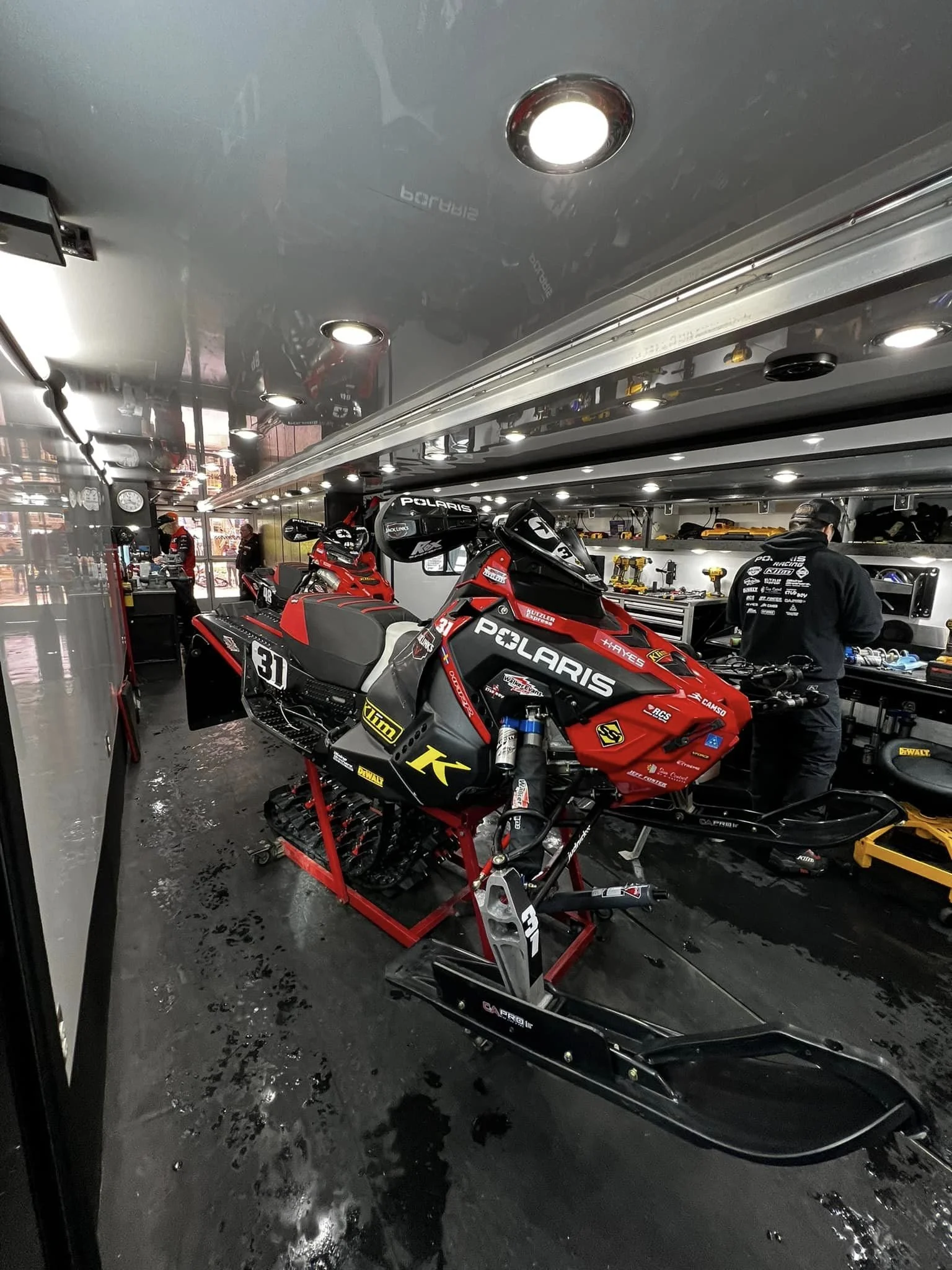 Indoor racing garage with a red and black Polaris snowmobile on a work stand, surrounded by tools and equipment, with several team members working in the background, under overhead lighting.