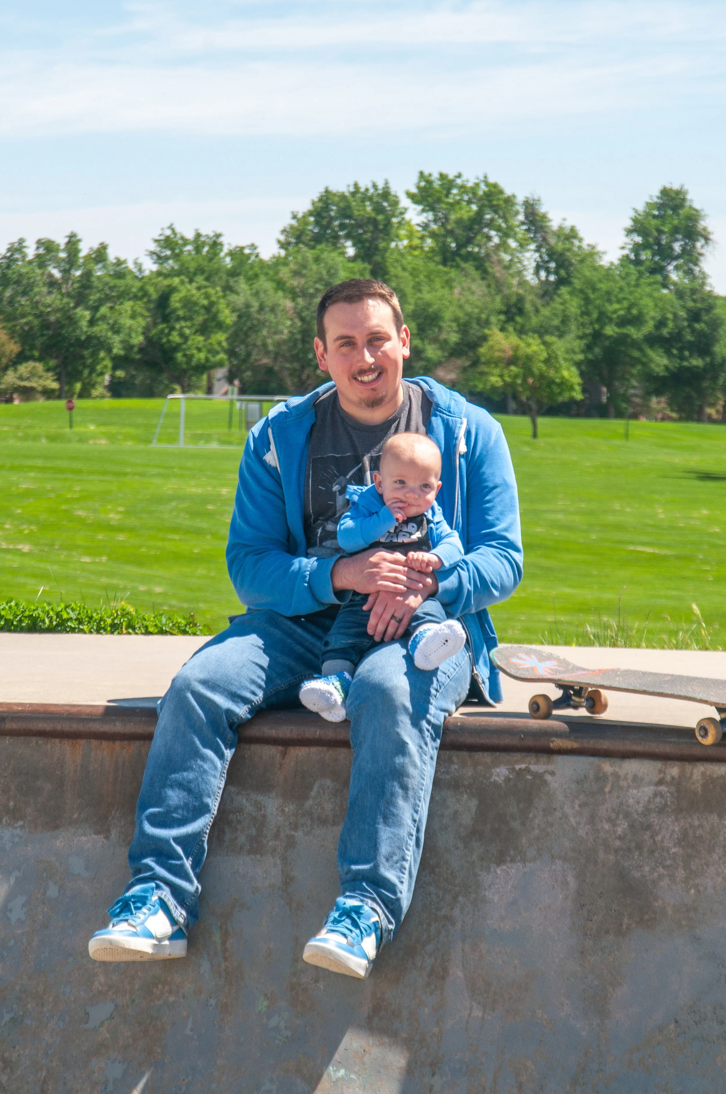 Man and baby sitting on the edge of a skate park with a skateboard nearby, green park and trees in the background, sunny sky.