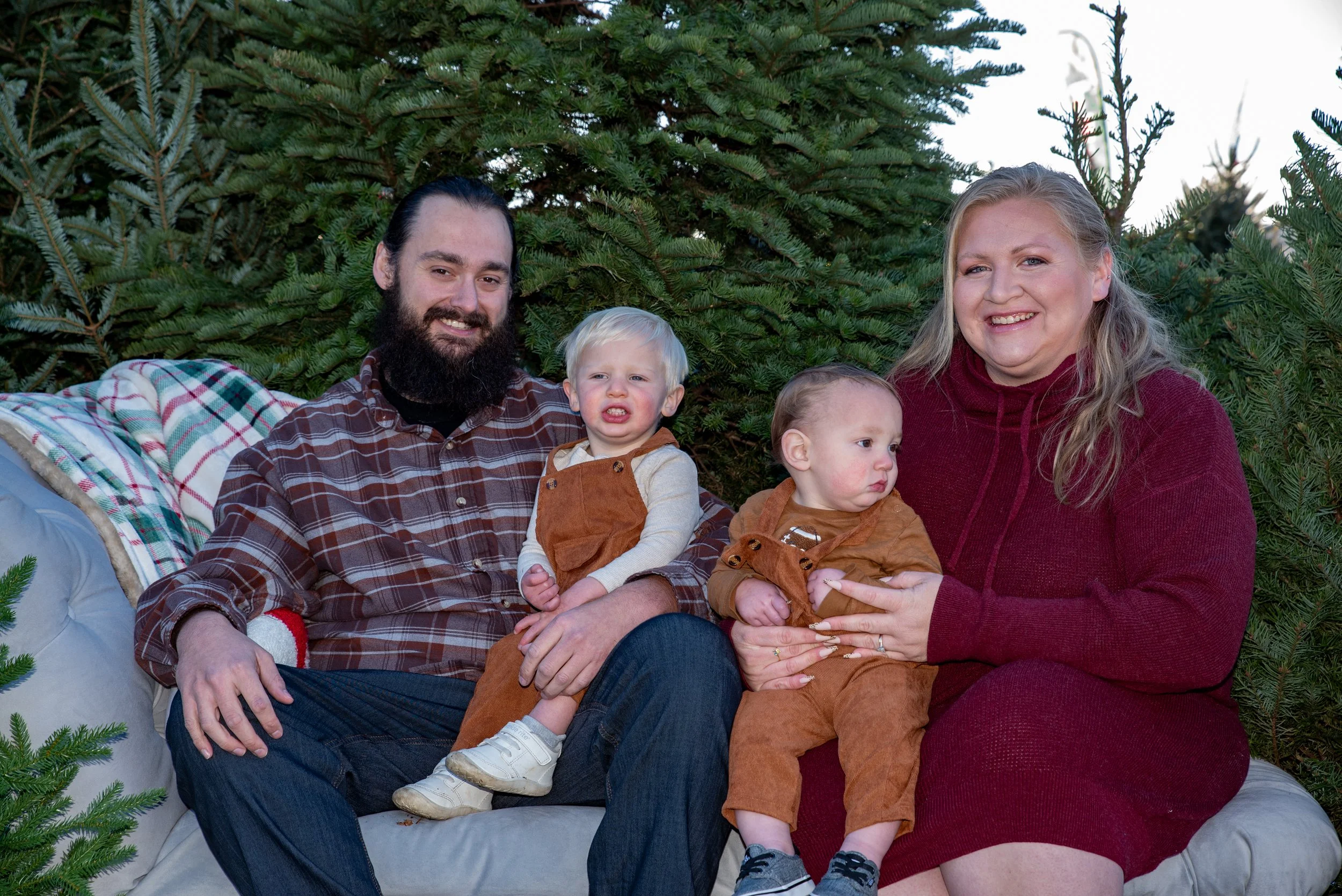 A family of four sitting on a sofa outdoors, surrounded by evergreen trees. The father has a beard and wears a plaid shirt, the mother has blonde hair and wears a red dress. The two young boys, both in brown outfits, are sitting between them.