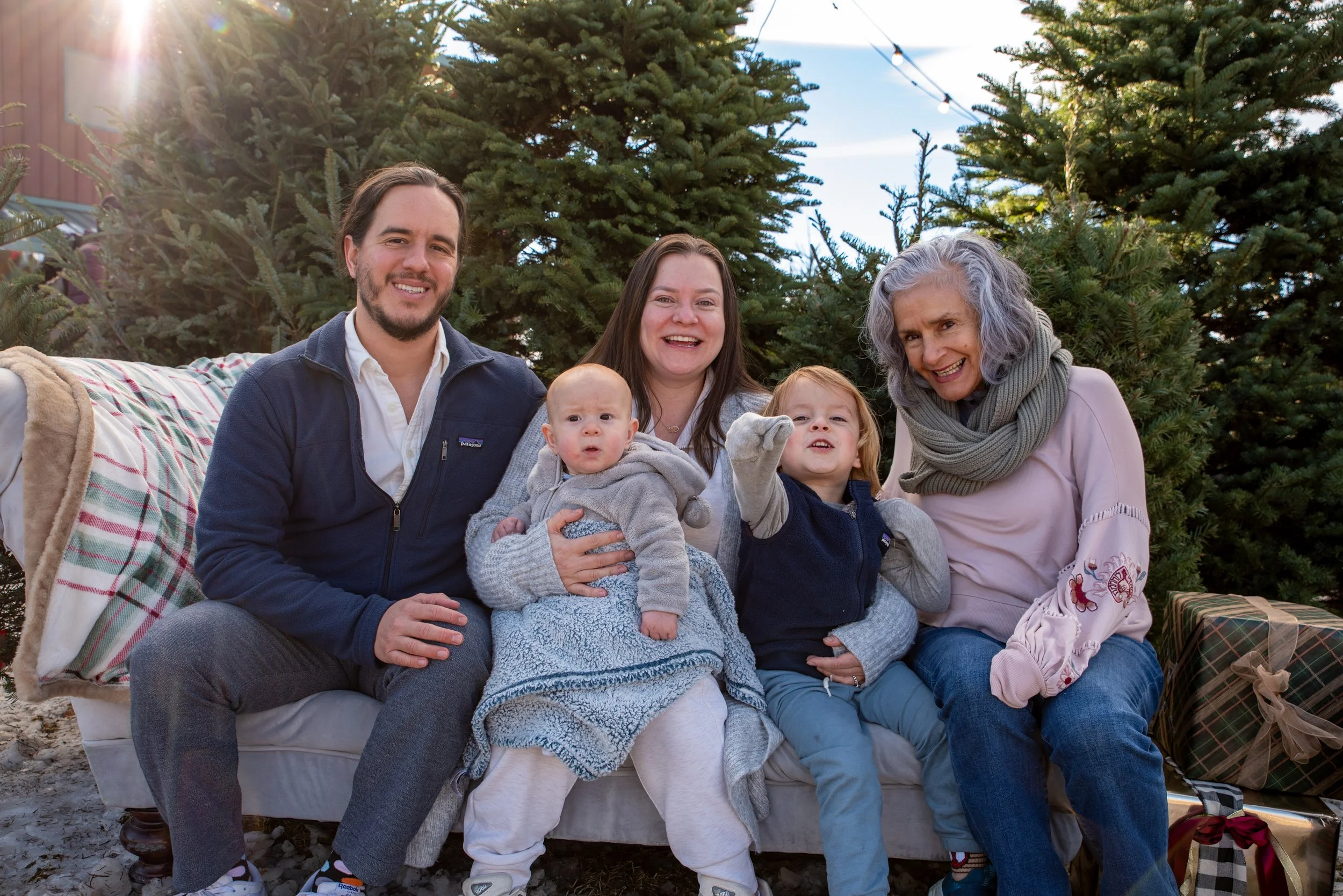Family of five, including three children, sitting outdoors near Christmas trees, smiling and enjoying a holiday gathering with presents nearby.