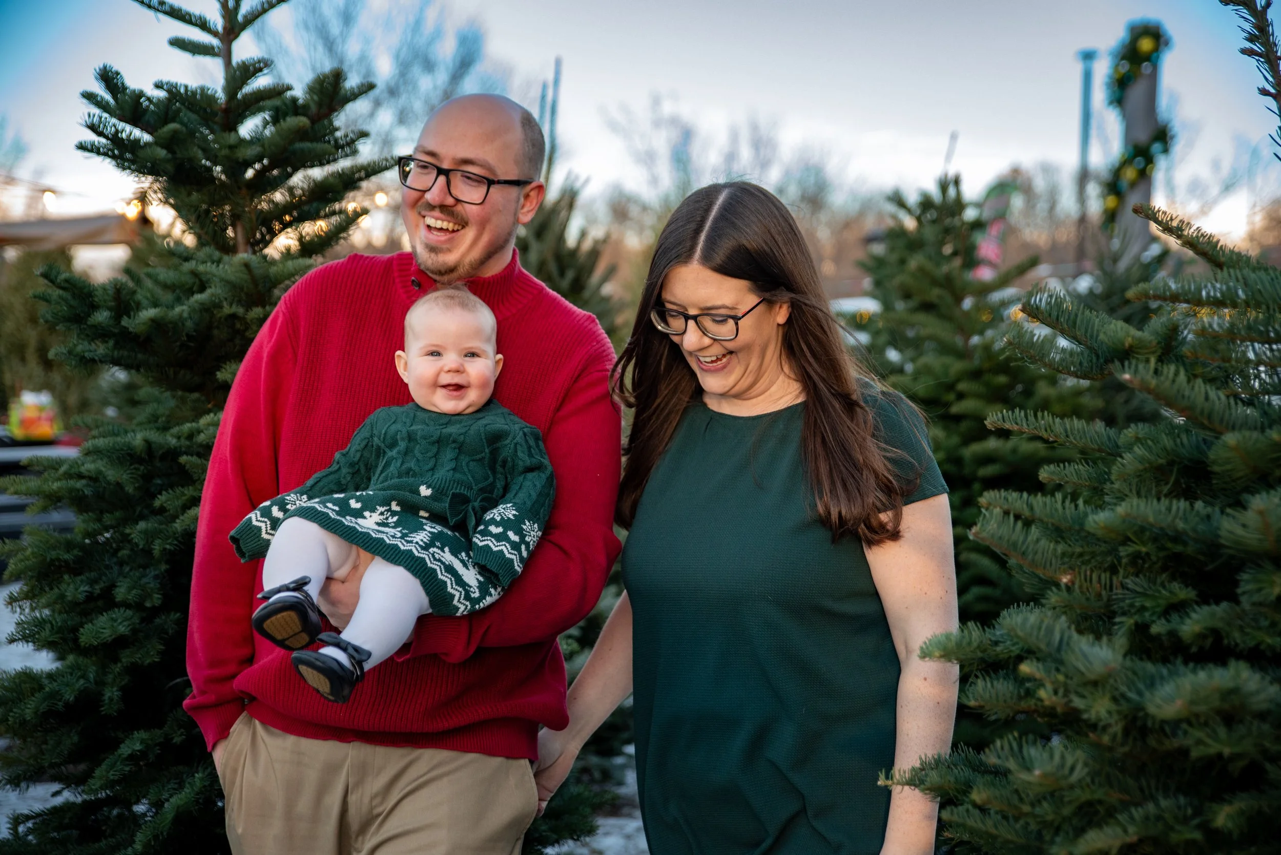 A happy family of three standing outdoors among Christmas trees, with a man in a red sweater holding a smiling baby girl in a festive green dress, and a woman in a dark green dress looking down and smiling.