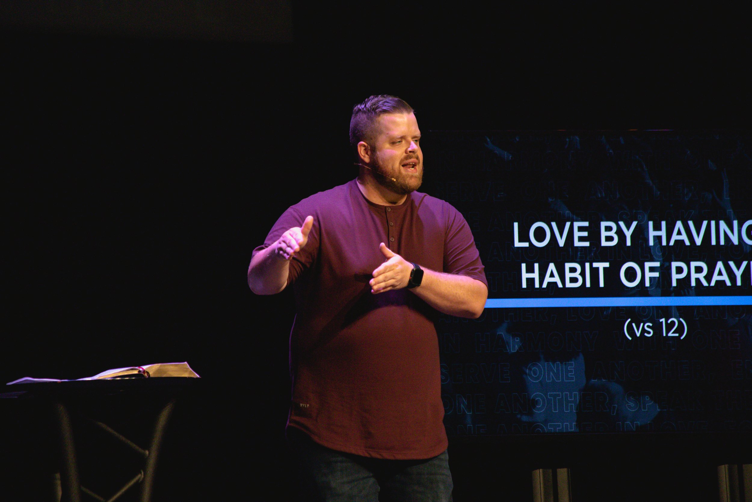 A man with a beard and short hair speaking on stage, gesturing with his hands, with text on a screen behind him that reads 'LOVE BY HAVING A HABIT OF PRAY'.