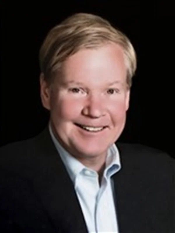 Portrait of a smiling man with light skin, blonde hair, wearing a dark blazer and a light blue collared shirt, against a black background.