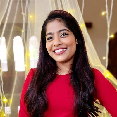 Young woman with long dark hair wearing a red top, smiling in front of a decorated background with white drapes and string lights.