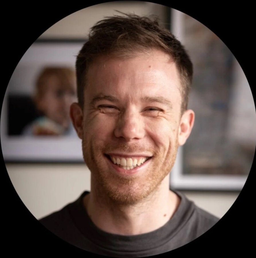 A smiling man with short brown hair and a beard, wearing a dark t-shirt, in a room with framed photos on the wall in the background.