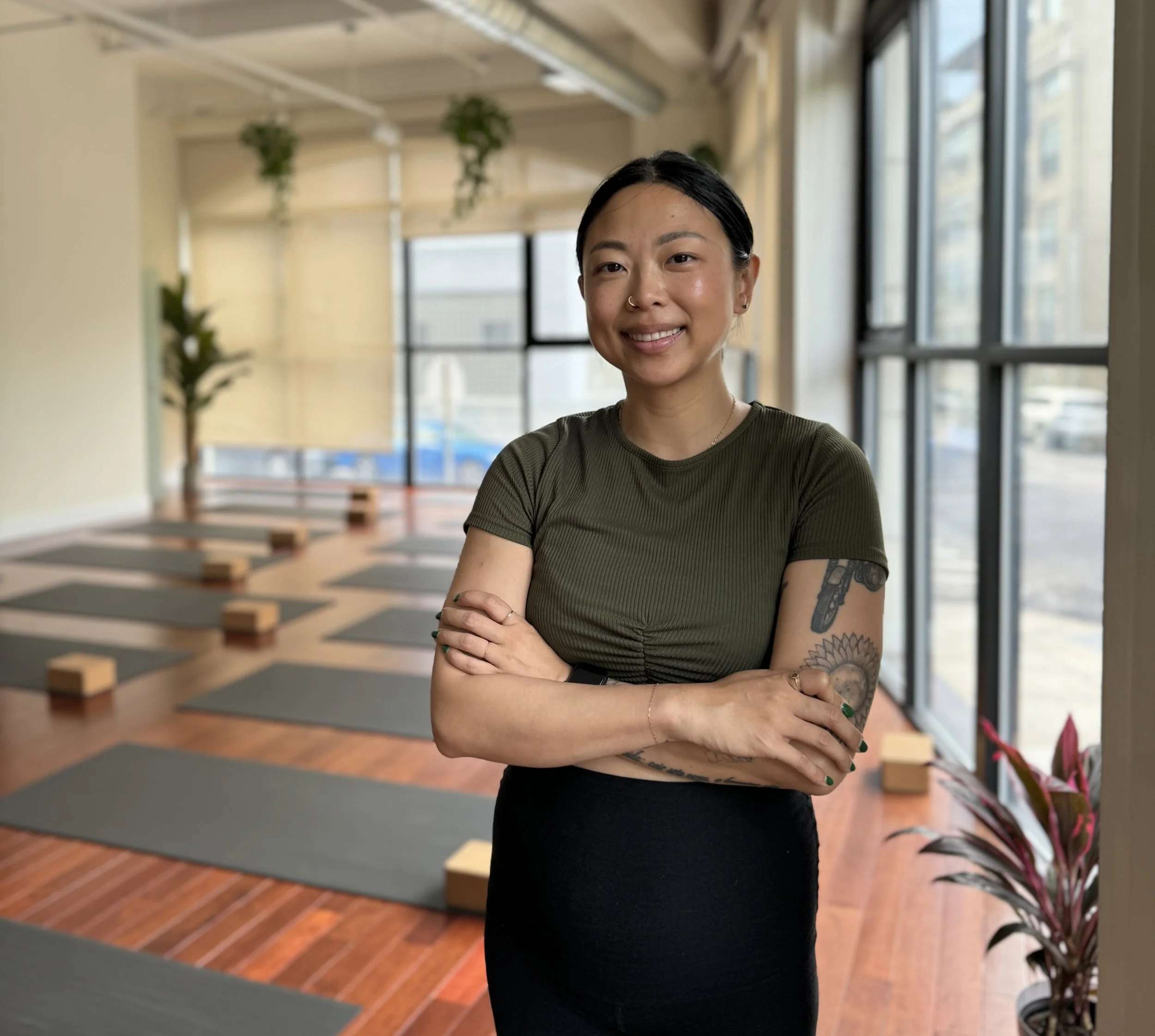 A smiling woman with tattoos, standing arms crossed inside a yoga studio with mats and blocks, large windows, and plants.