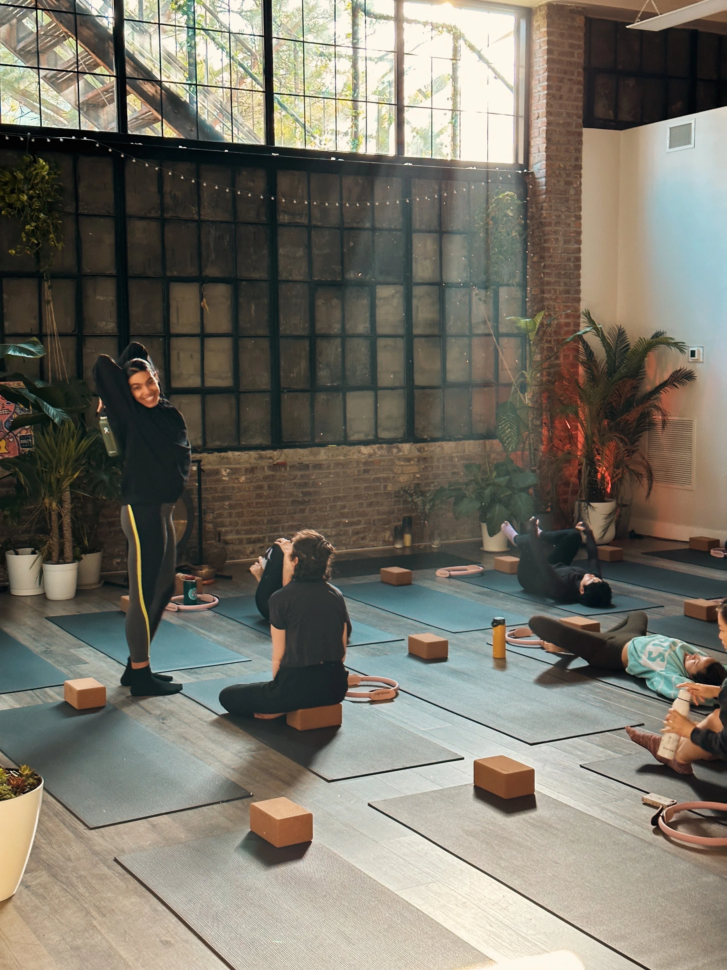 A yoga class in a rustic studio with large windows, plants, and participants lying on mats while an instructor stands and smiles.