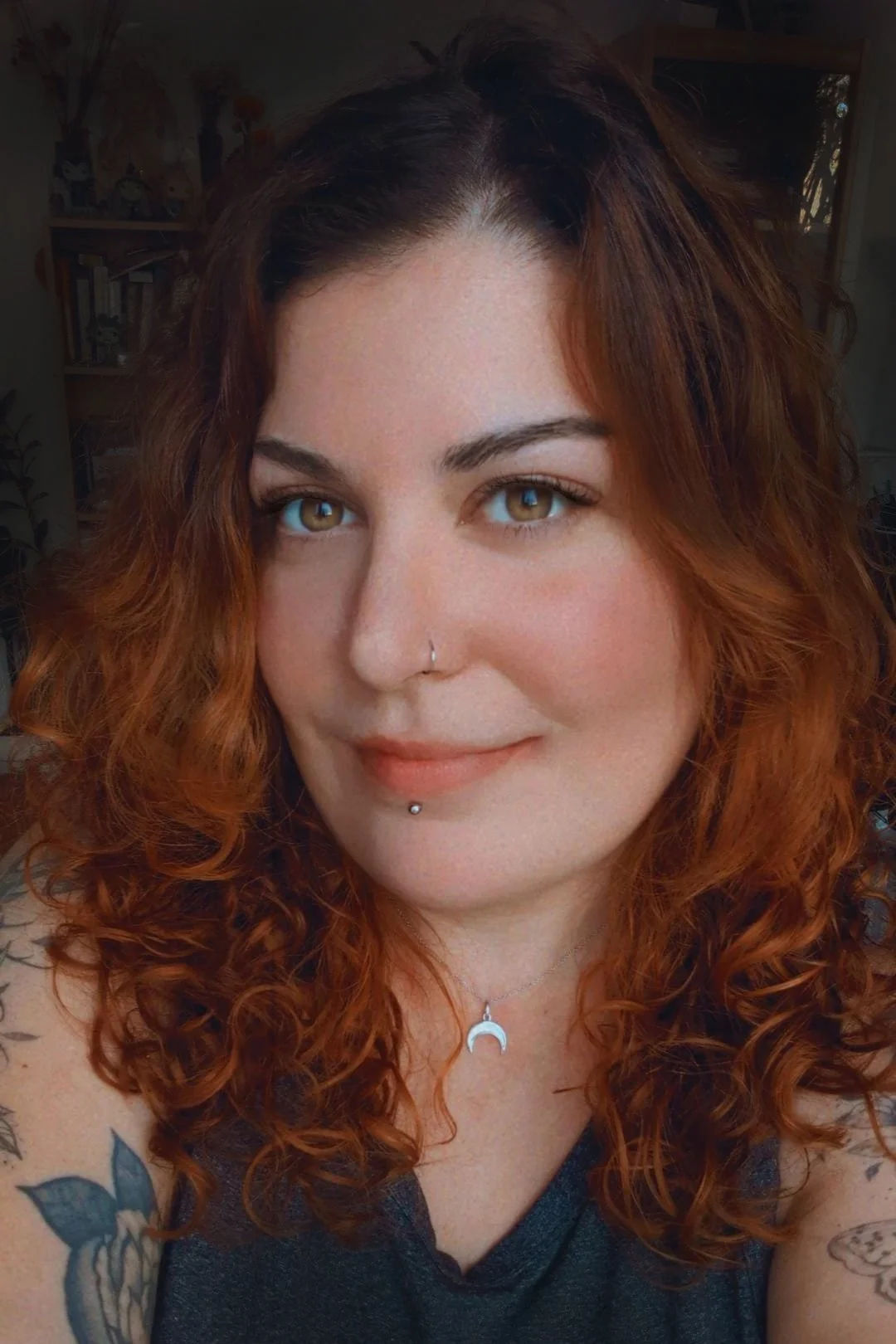 Femme aux cheveux bouclés roux portant un piercing au nez et au labret, avec un collier en forme de lune, fond flou.