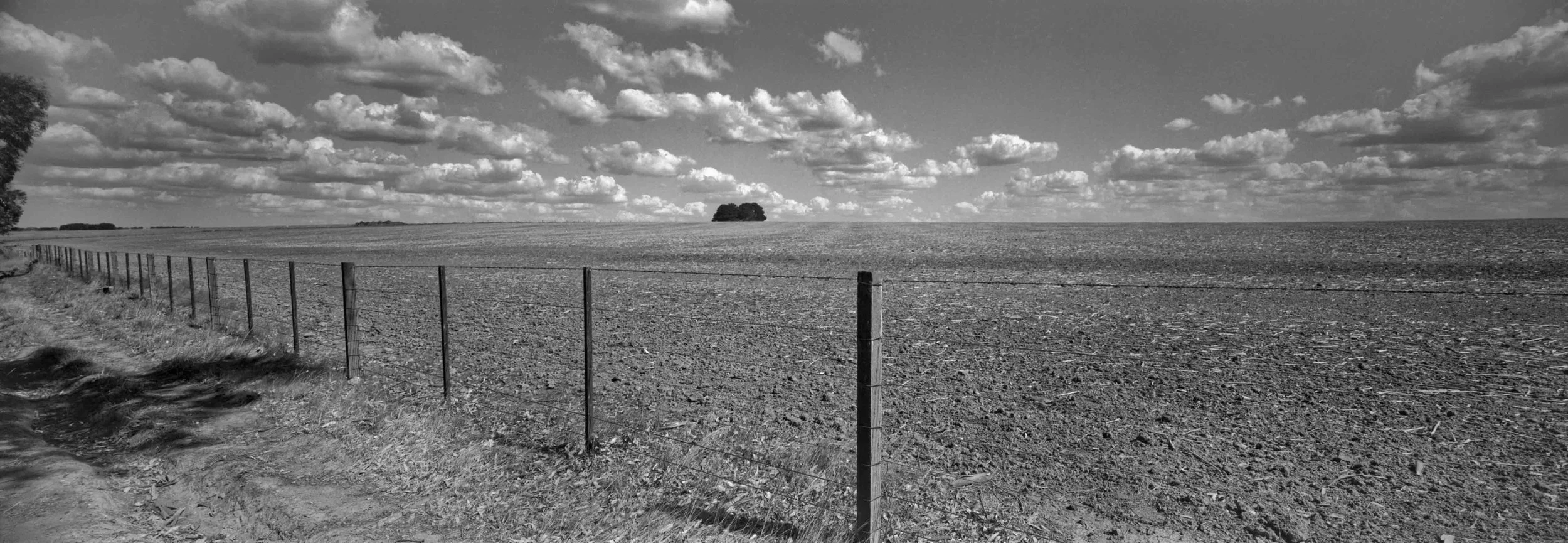 A black and white photo of a rural landscape featuring wide fields, a fence line along the dirt roadside, distant trees, and clouds in the sky.