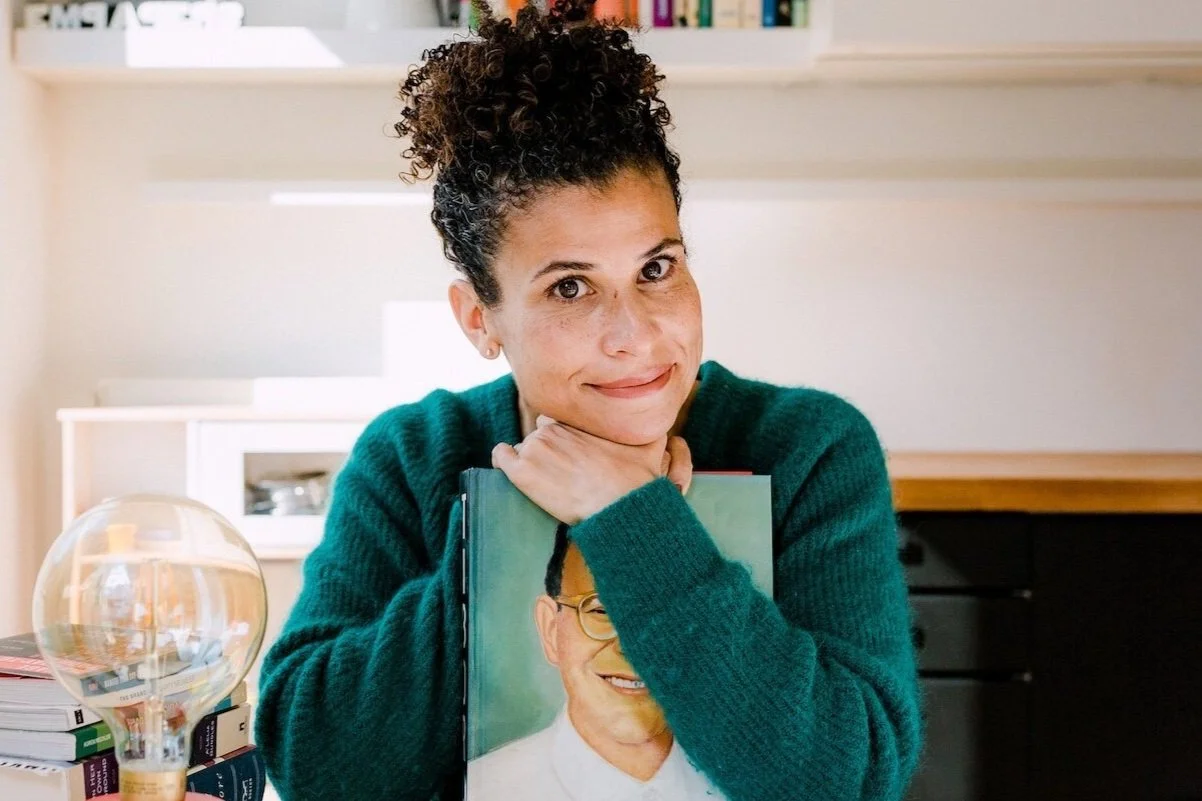 A woman with curly hair in a bun, wearing a green sweater, is sitting at a table holding a book with a man's portrait on the cover. She is resting her chin on her hand, smiling gently at the camera. There are stacks of books and a round lamp on the table. A bookshelf with books is visible in the background.