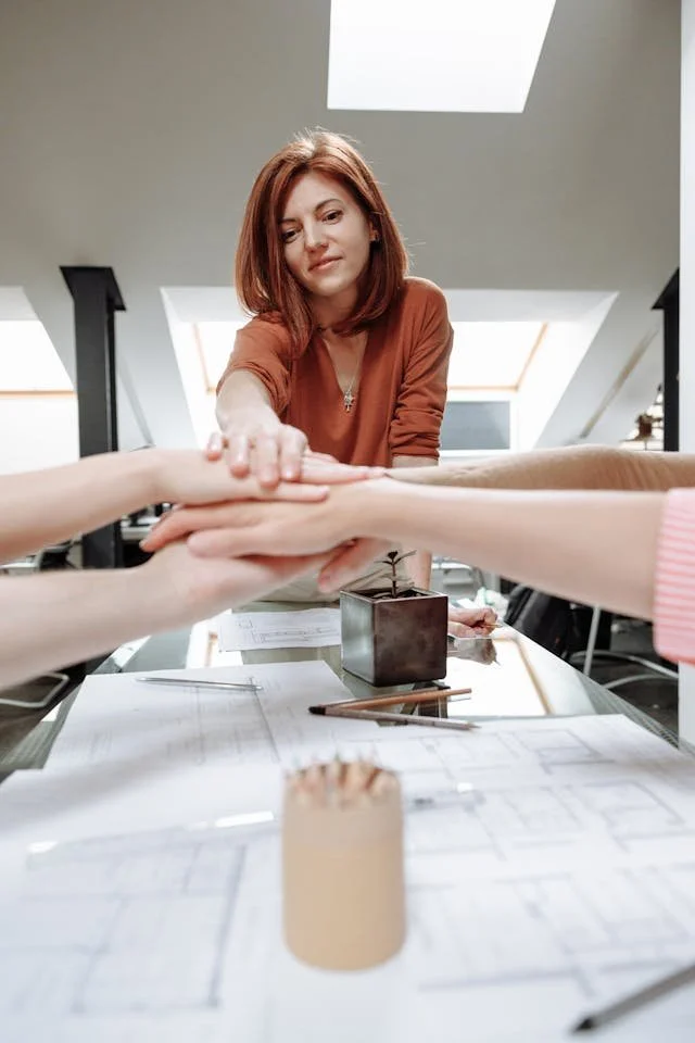 A woman with red hair appears to be guiding or coaching two other individuals, as their hands are stacked together in a gesture of teamwork or support, with architectural plans and writing tools on the table.