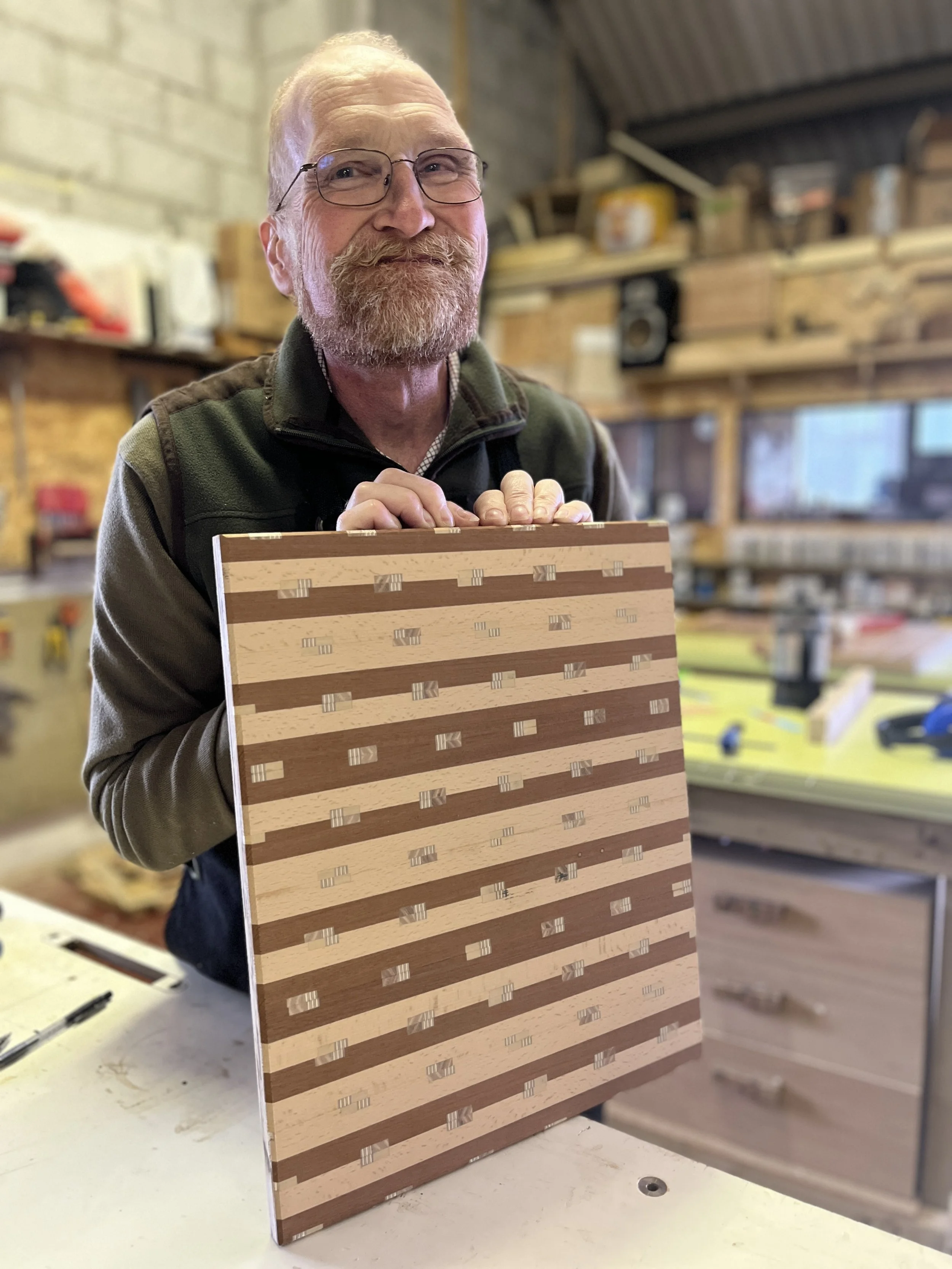 A man with glasses and a beard smiling and holding a piece of wood with horizontal striped patterns in a workshop.