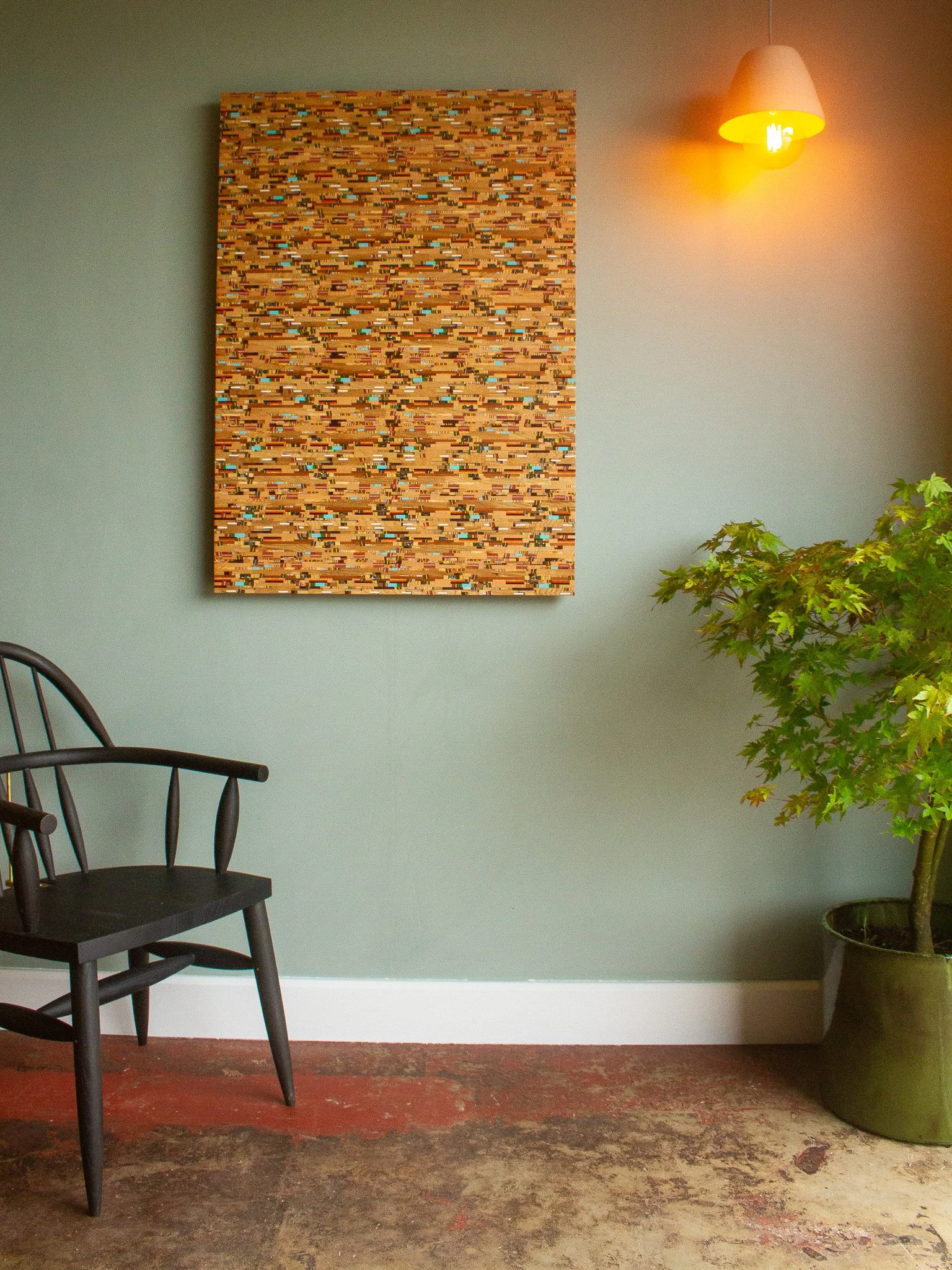 Interior of a room with a black wooden chair on the left, a large abstract wooden wall art piece on a light green wall, a yellow wall lamp turned on, and a potted green leafy plant on the right.