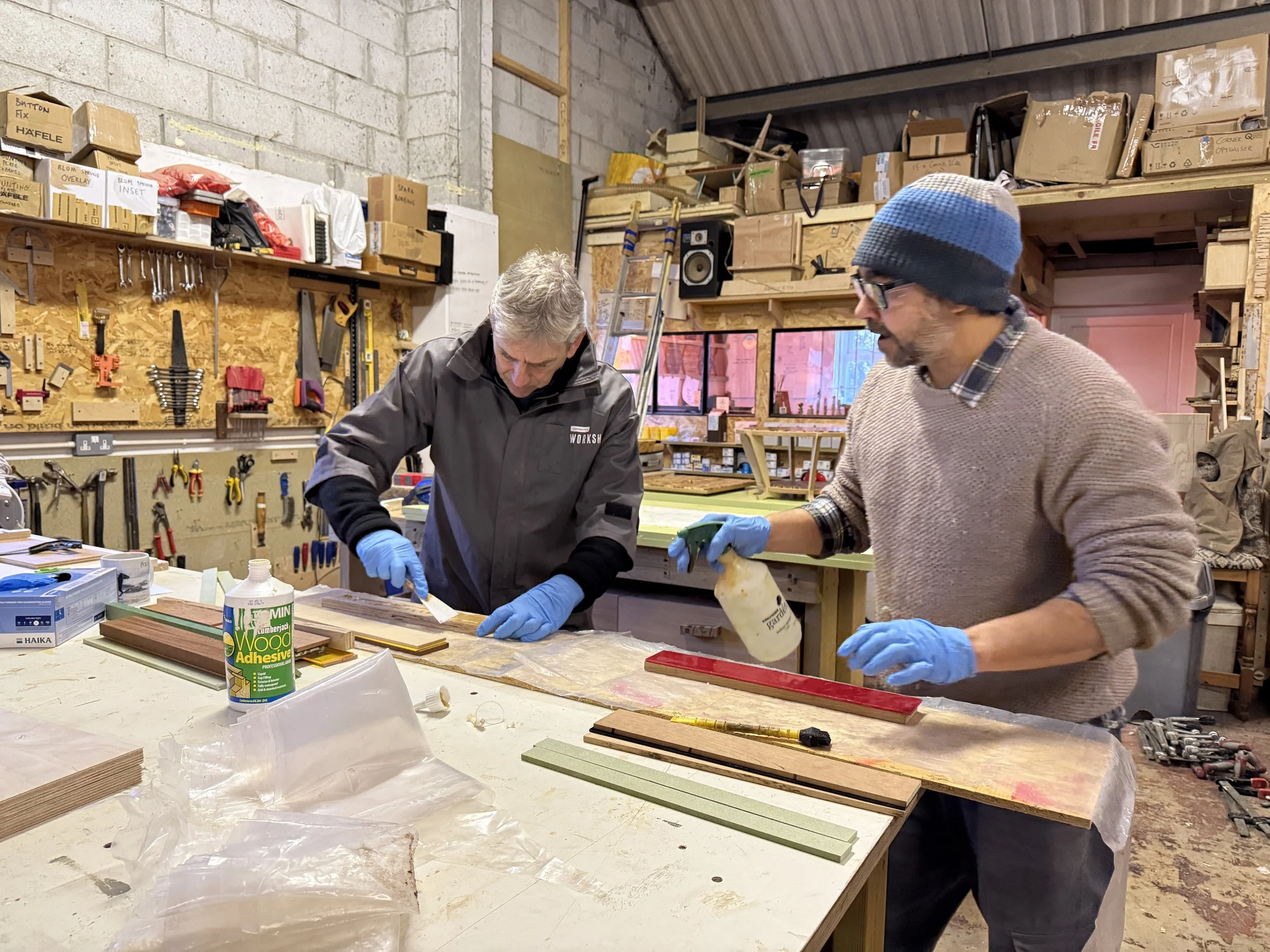 Two men working together in a woodworking workshop, wearing gloves, surrounded by tools, wood, and supplies.