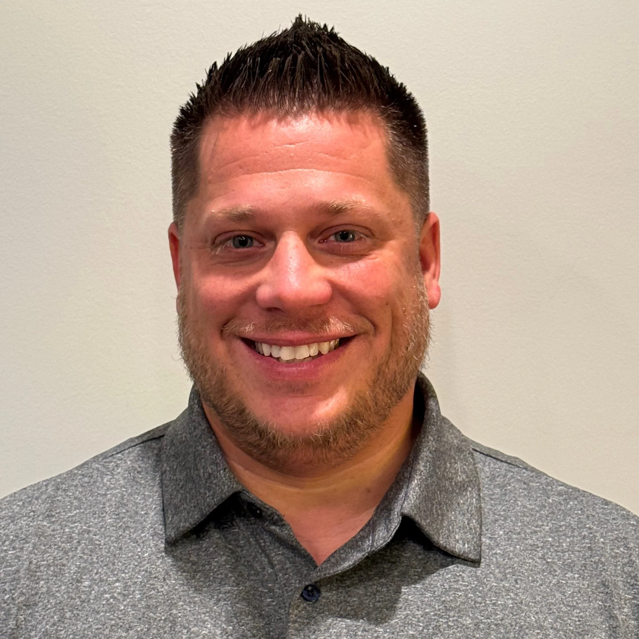Close-up of a smiling man with short dark hair, wearing a gray textured blazer and black shirt, standing against a plain light-colored wall.