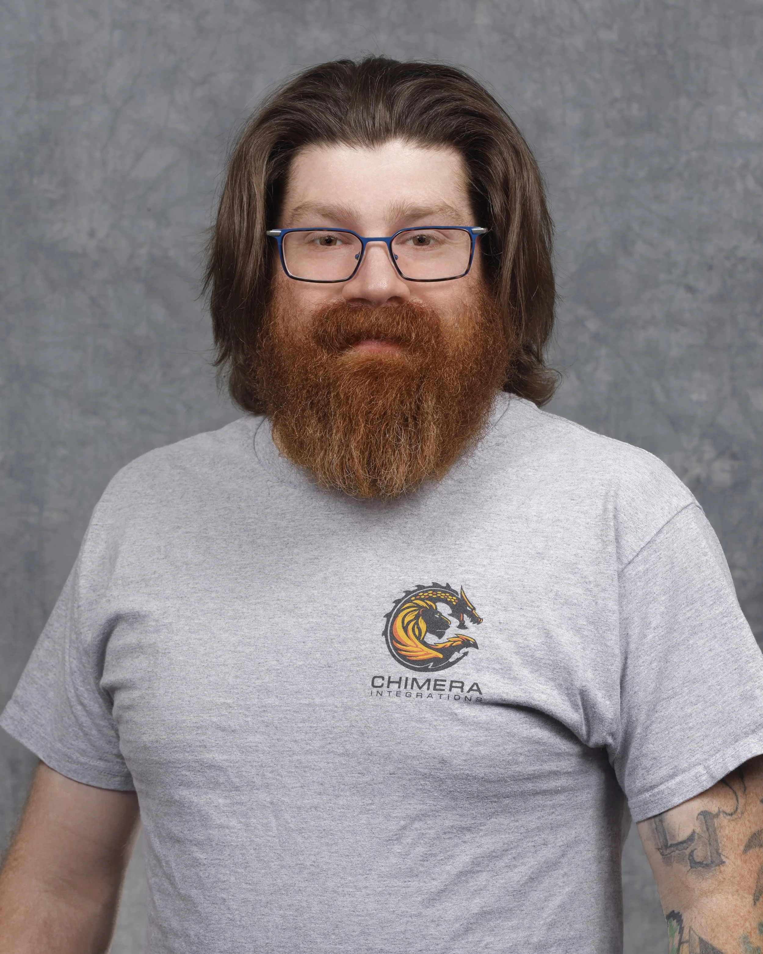 A man with dark hair, glasses, and a red beard, wearing a black T-shirt, standing in front of shelves filled with boxes and storage items.