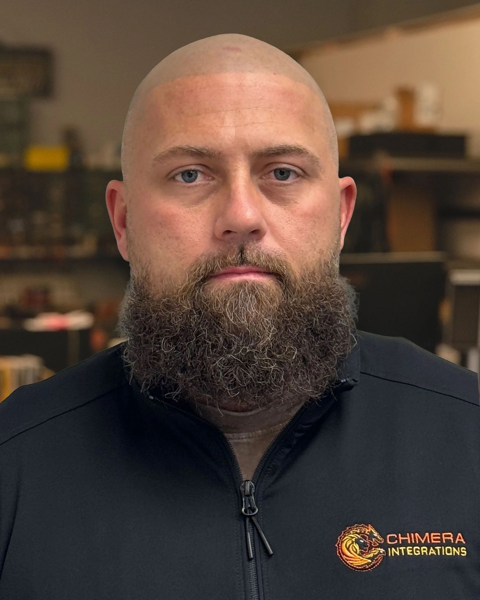 Close-up of a man with a shaved head and full beard, wearing a black jacket with a logo on the chest that reads "Chimera Integrations" and an image of a lion's head.