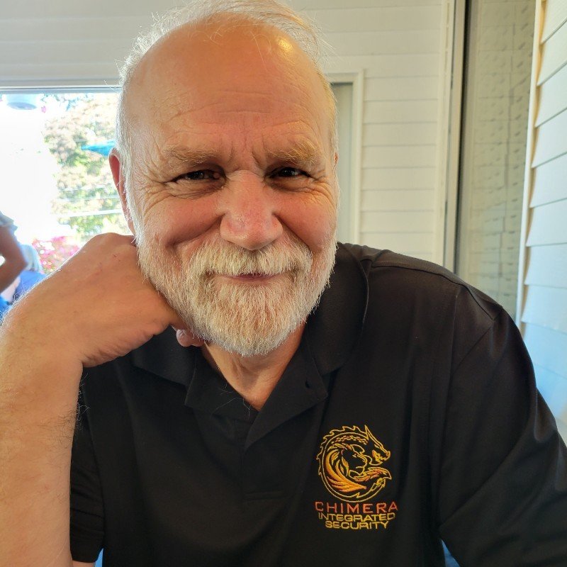 A smiling elderly man with a white beard and short white hair, wearing a black polo shirt with a logo that says 'Chimera Integrated Security' and depicts a lion's head in gold and orange.