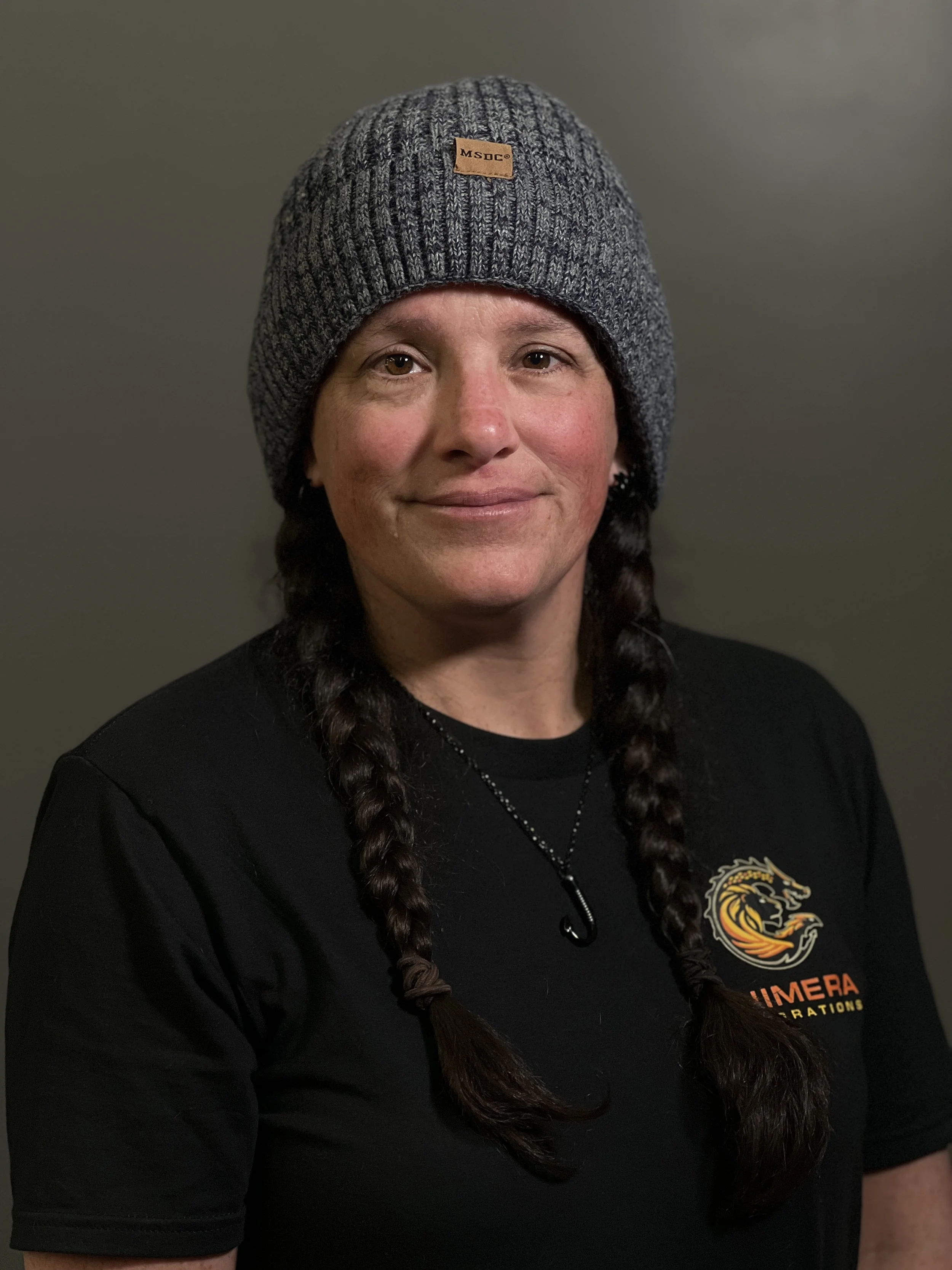 Woman with long braided hair wearing a knit gray beanie and a black T-shirt with a logo of a lion's head, text 'LIMERA' and 'OPERATIONS'.