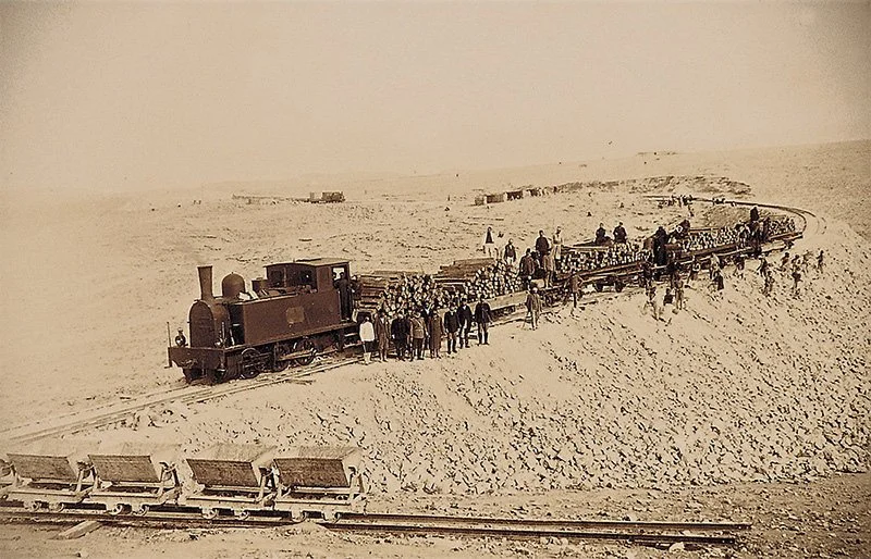 Historical black-and-white photograph of a crowded steam train in a desert landscape with many people on and around the train, and a train car with chairs in the foreground.
