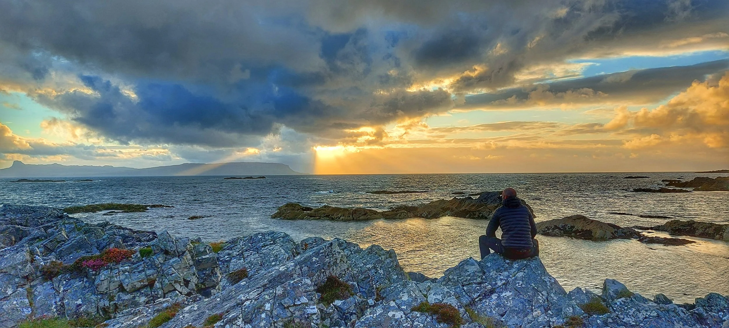 A person sitting on rocks by the ocean watching a sunset with dark clouds and distant mountains in the background.