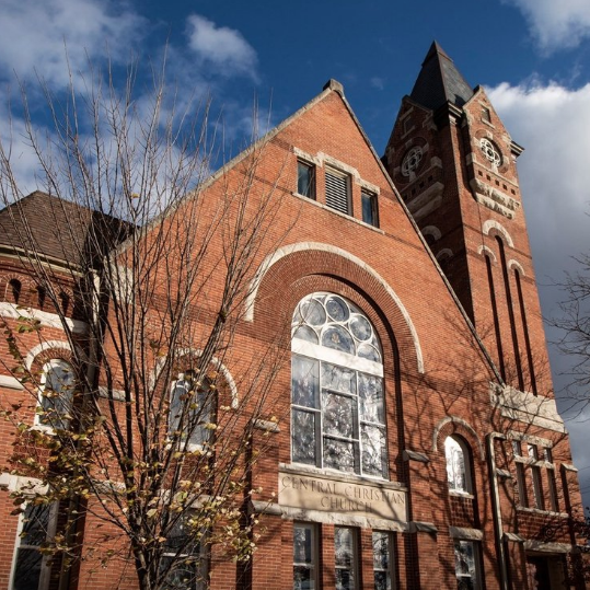 Historic red brick church with tall steeple, arched windows, and leafless trees on a clear day with clouds.