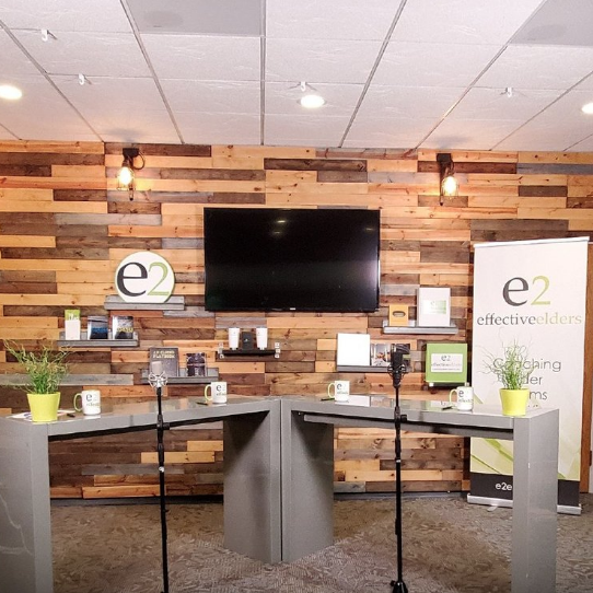 A modern office space with a wooden accent wall featuring a mounted TV screen and various brochures. There are two podiums with microphones, green plants, and mugs. A standing banner displays the text 'effectiveelders.' The decor has a professional and inviting atmosphere.