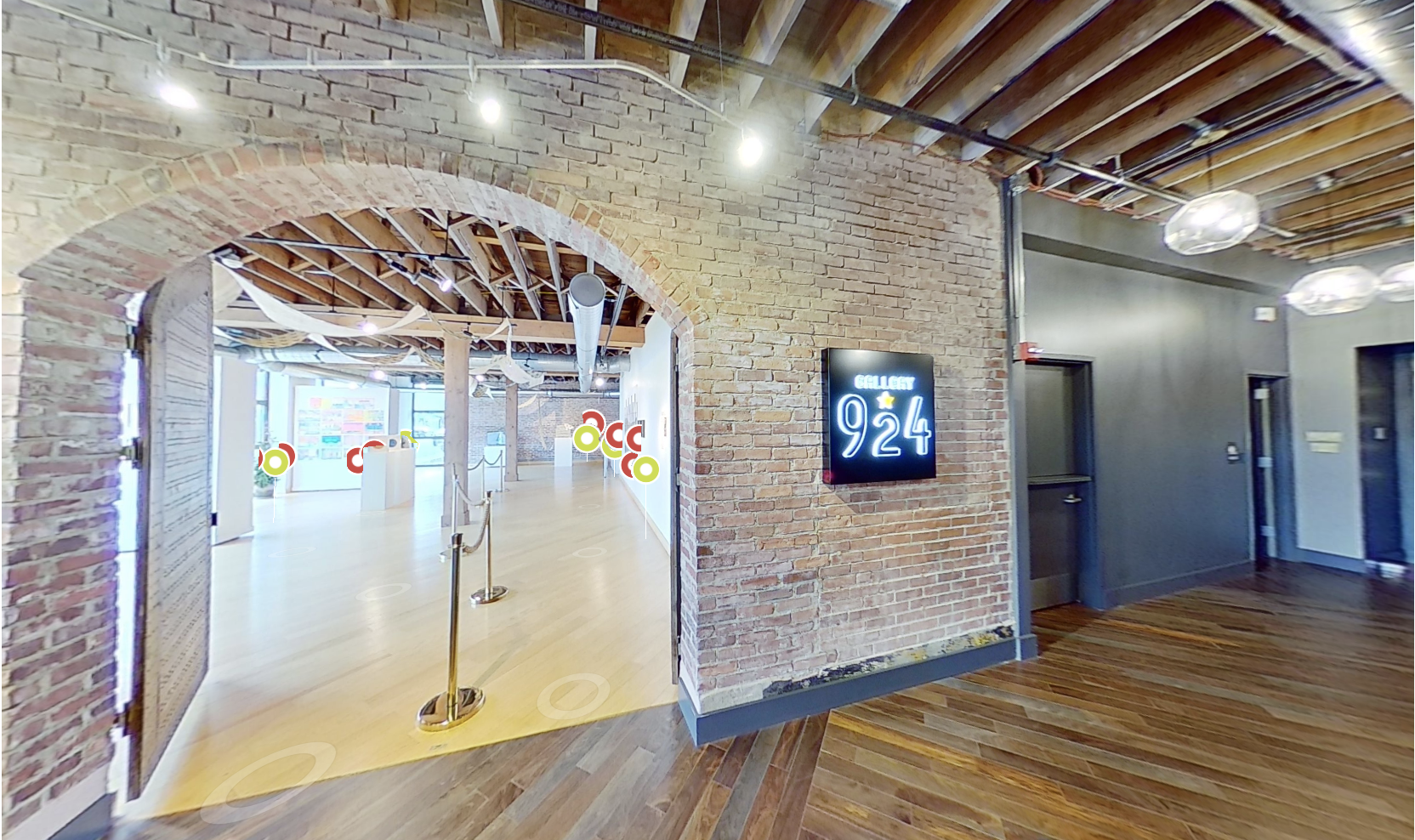 Interior of Gallery 924 with exposed brick walls, wooden floors, and a LED sign on the wall. Open space with modern lighting and ductwork, featuring an entrance to an exhibit area.