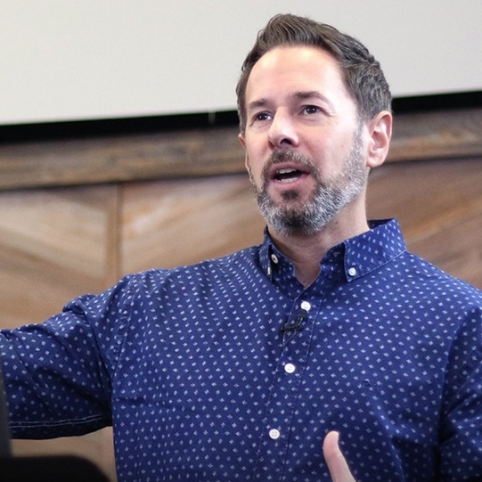 A man speaking, wearing a blue patterned shirt, in a professional setting.