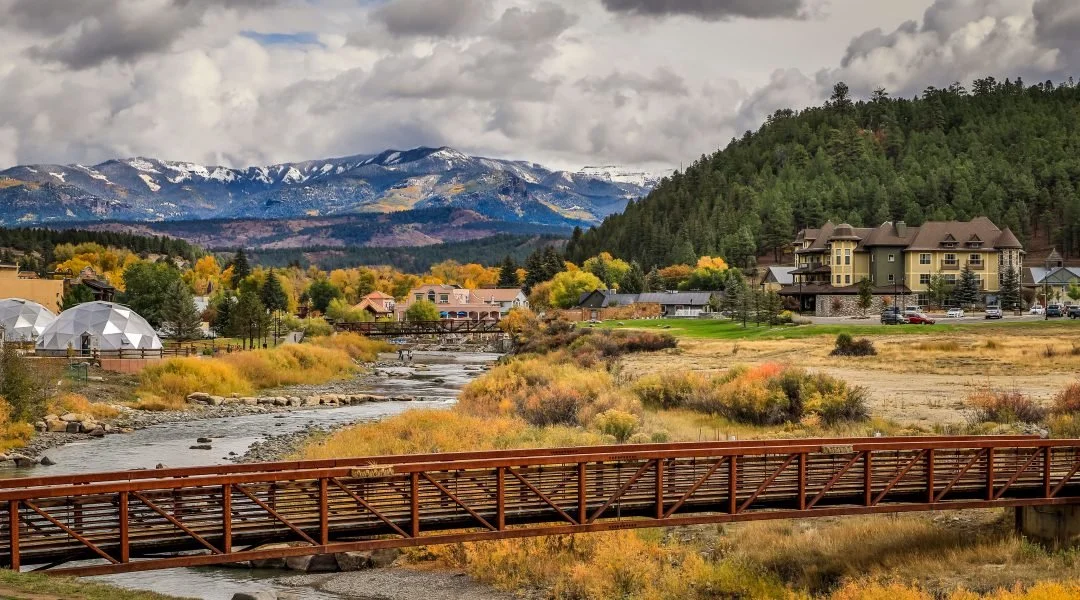 A scenic landscape featuring a river, colorful autumn foliage, a wooden bridge, residential buildings, and snow-capped mountains in the background.