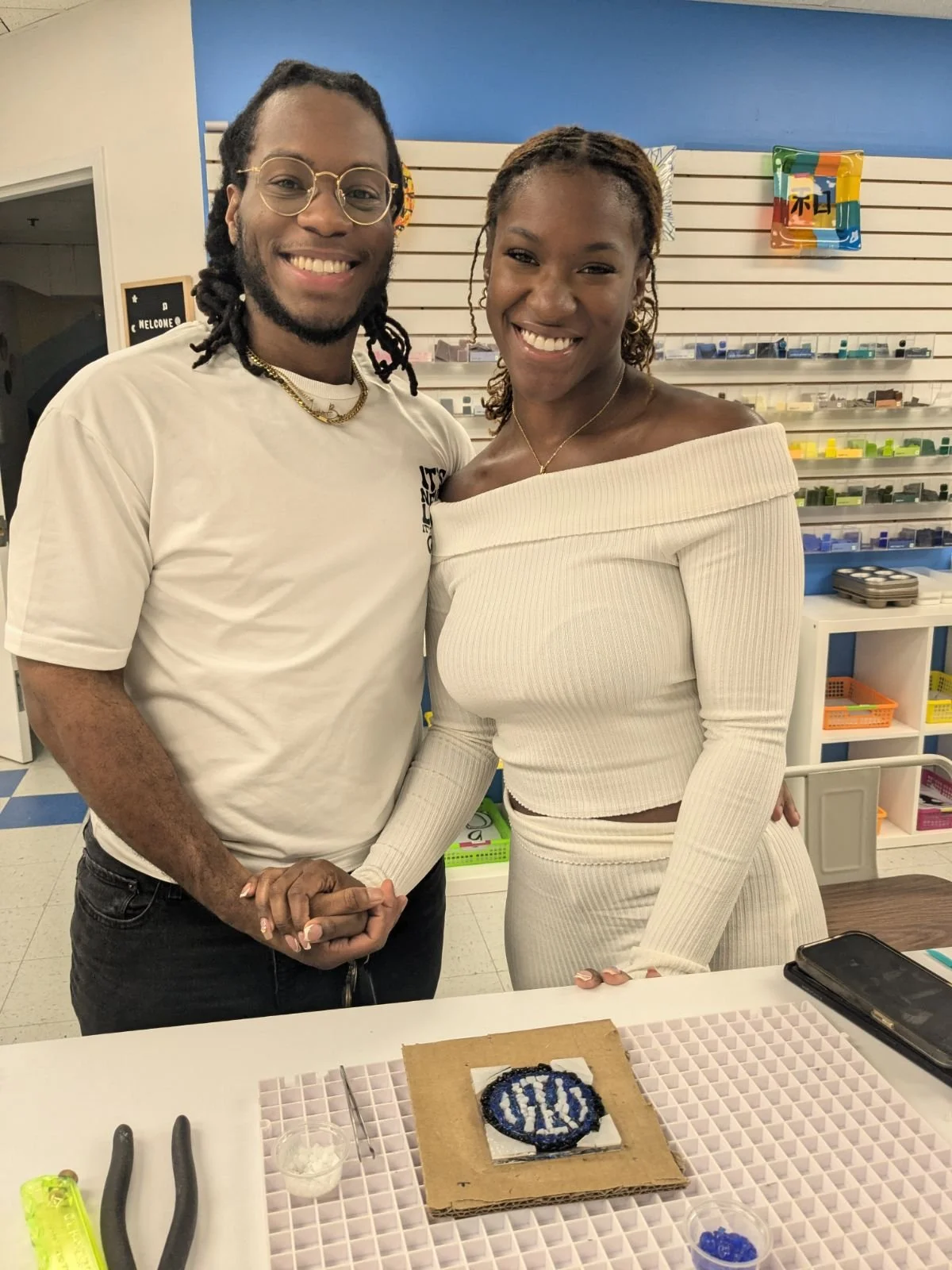 A smiling man and woman holding hands in a craft workshop. The man has glasses and dreadlocks, the woman has curly hair. A craft project on the table features a circular beaded design with the word 'LOVE'.