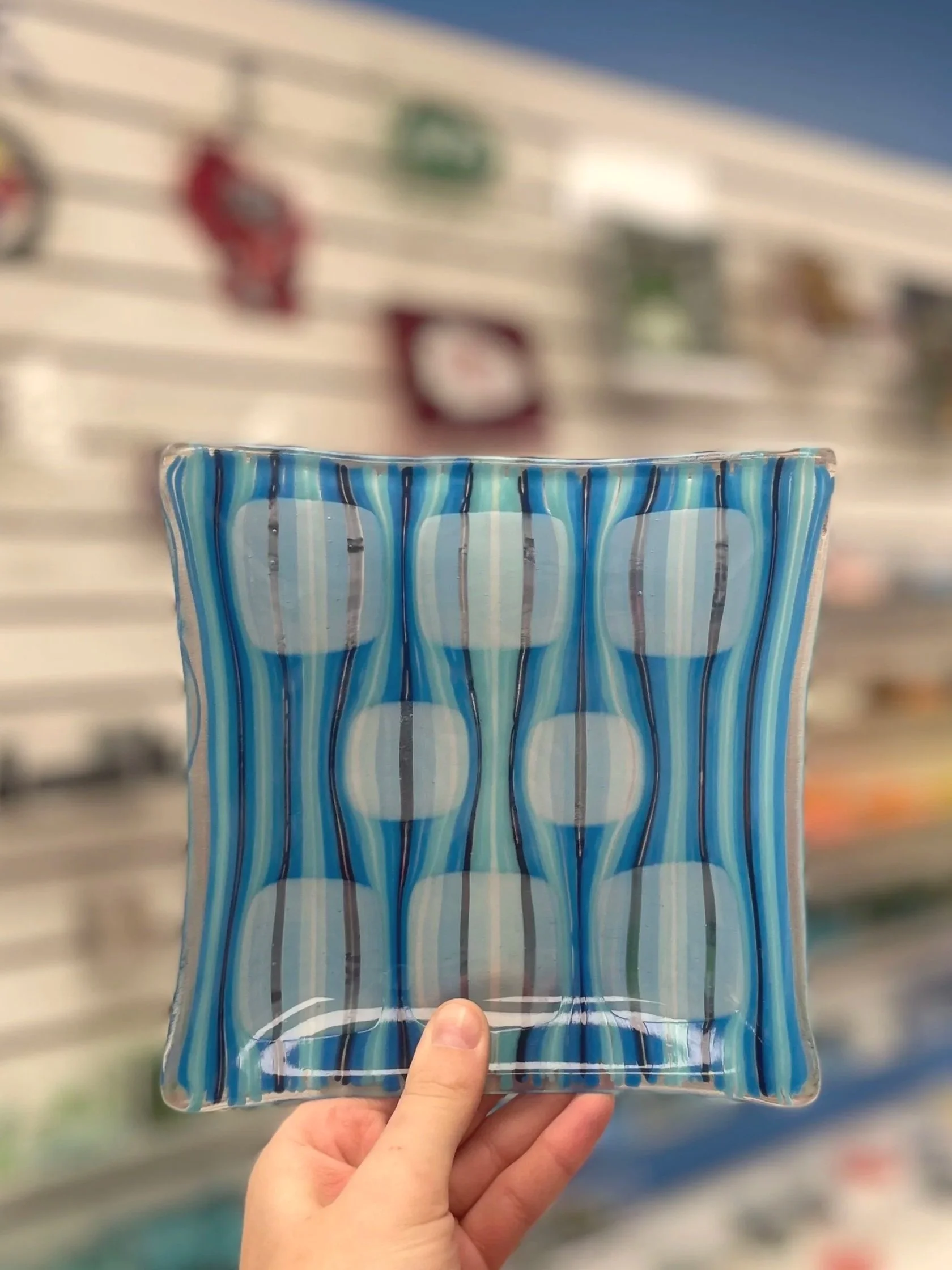 A person holding a square, glass plate with a blue and white striped pattern in a store with shelves in the background.