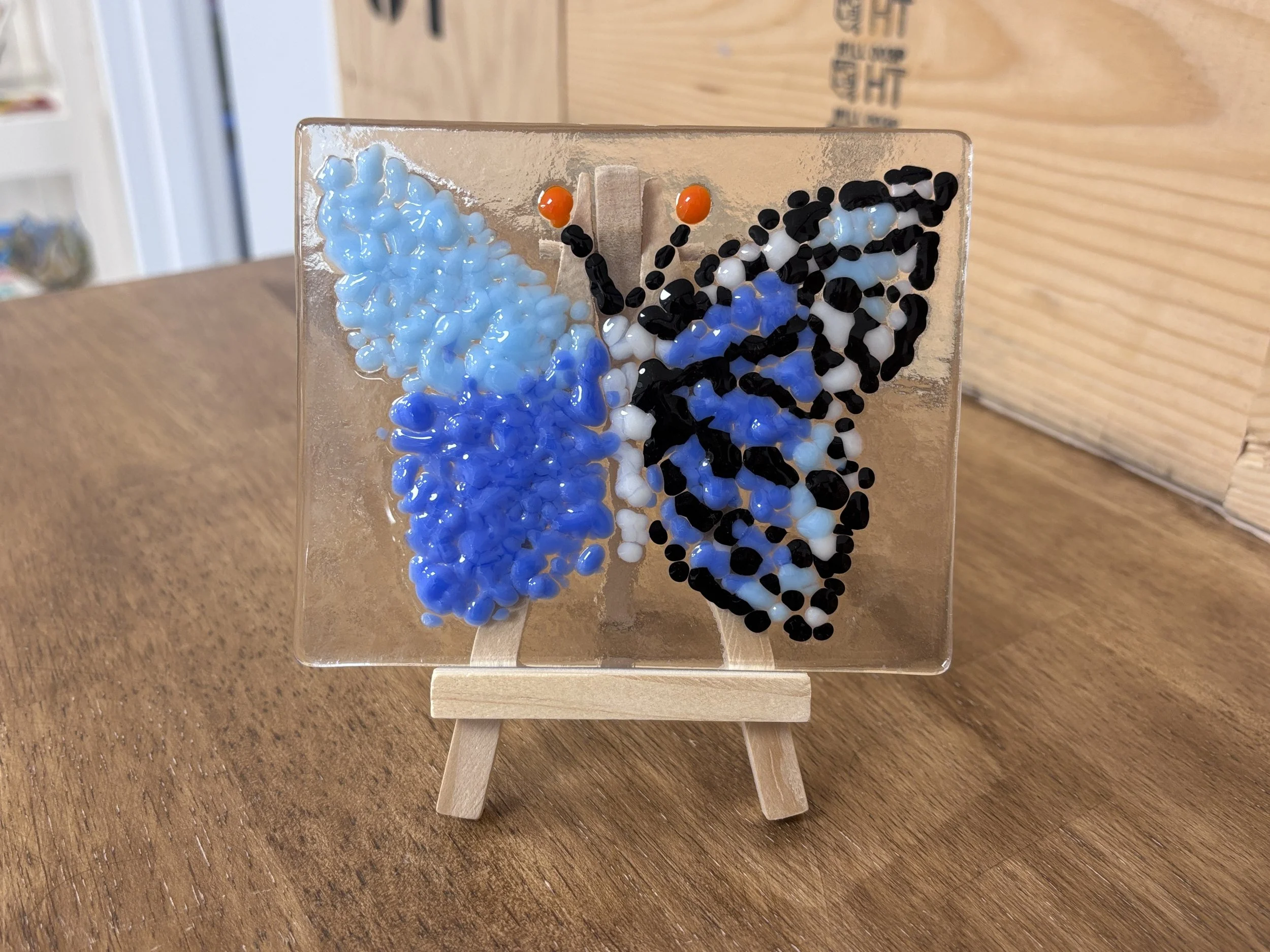Colorful mosaic artwork on a small easel featuring clouds, water, grass, and trees made with small beads or stones.