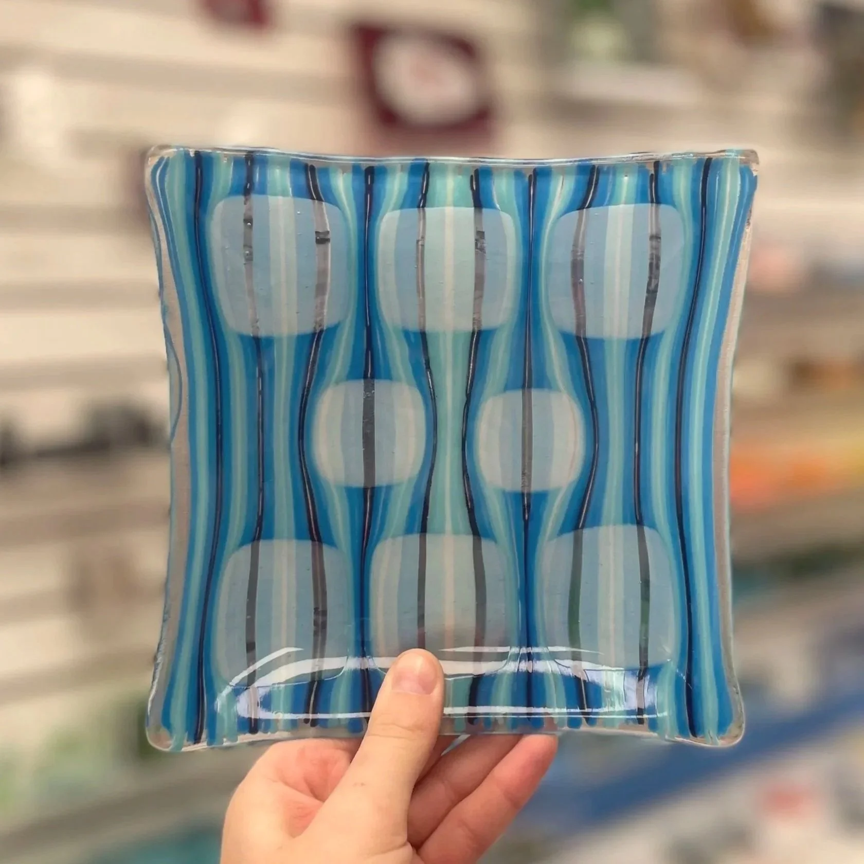 A person holding a square, glass plate with a blue and white striped pattern in a store with shelves in the background.