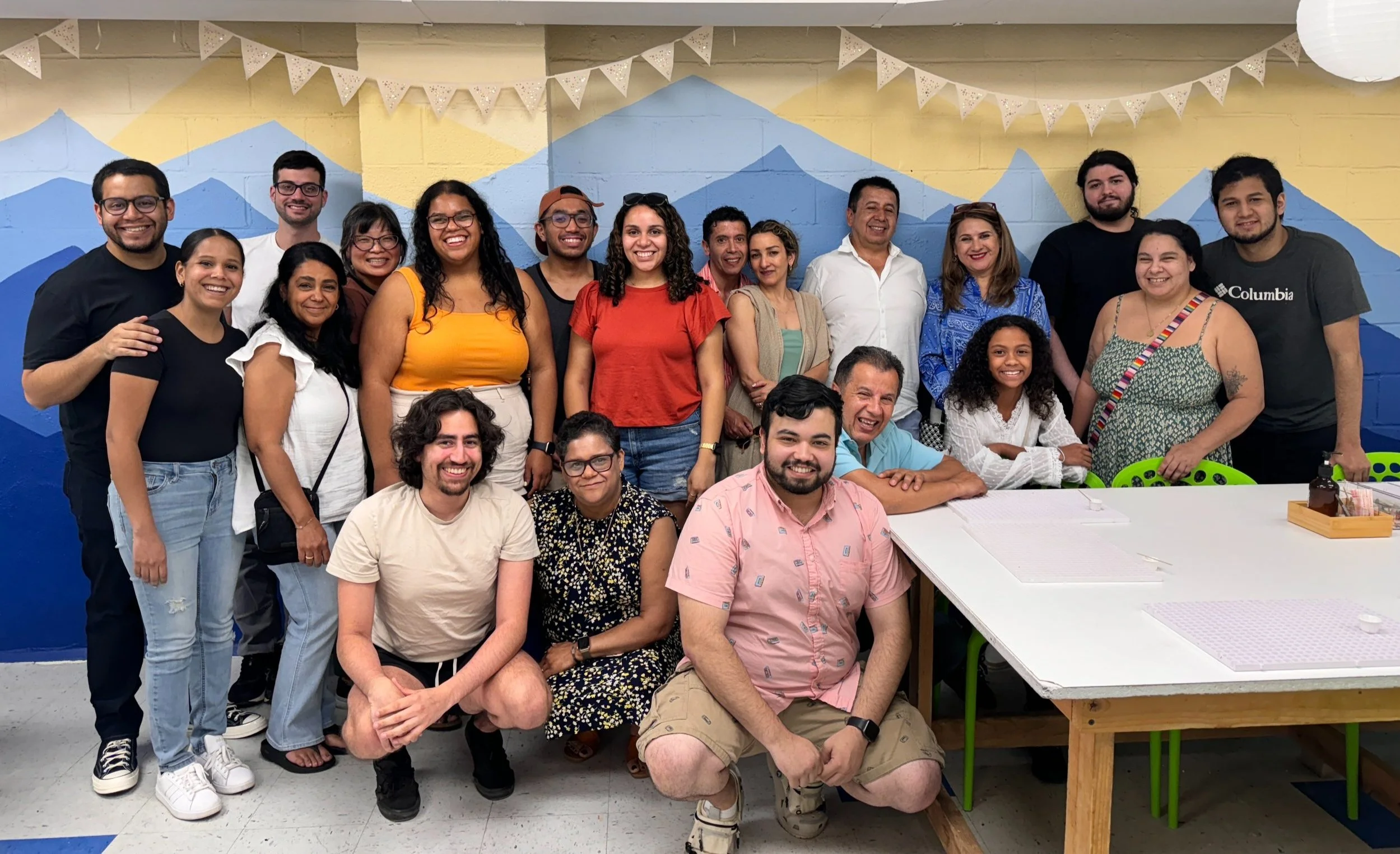 Group of people smiling and posing for a photo in an indoor space with a colorful mountain mural and decorative banners on the wall.