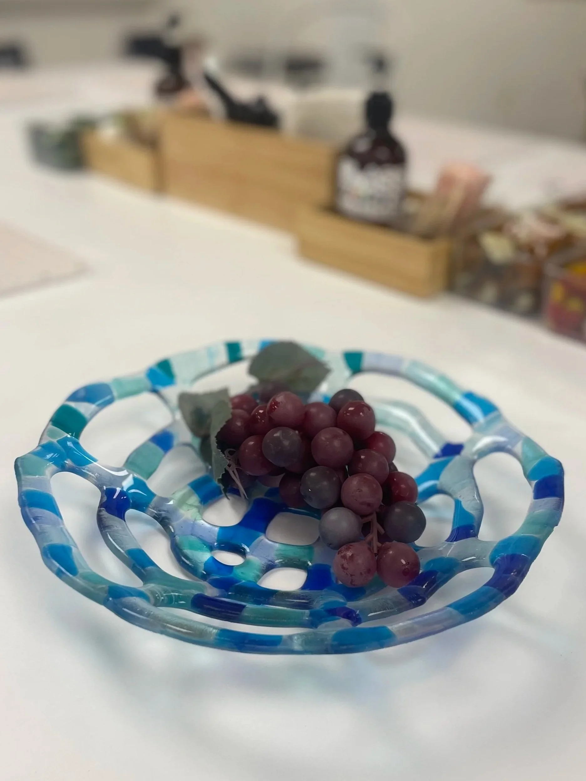 A glass bowl with blue and light blue streaks containing a bunch of red and purple grapes, on a white surface with a blurred background.
