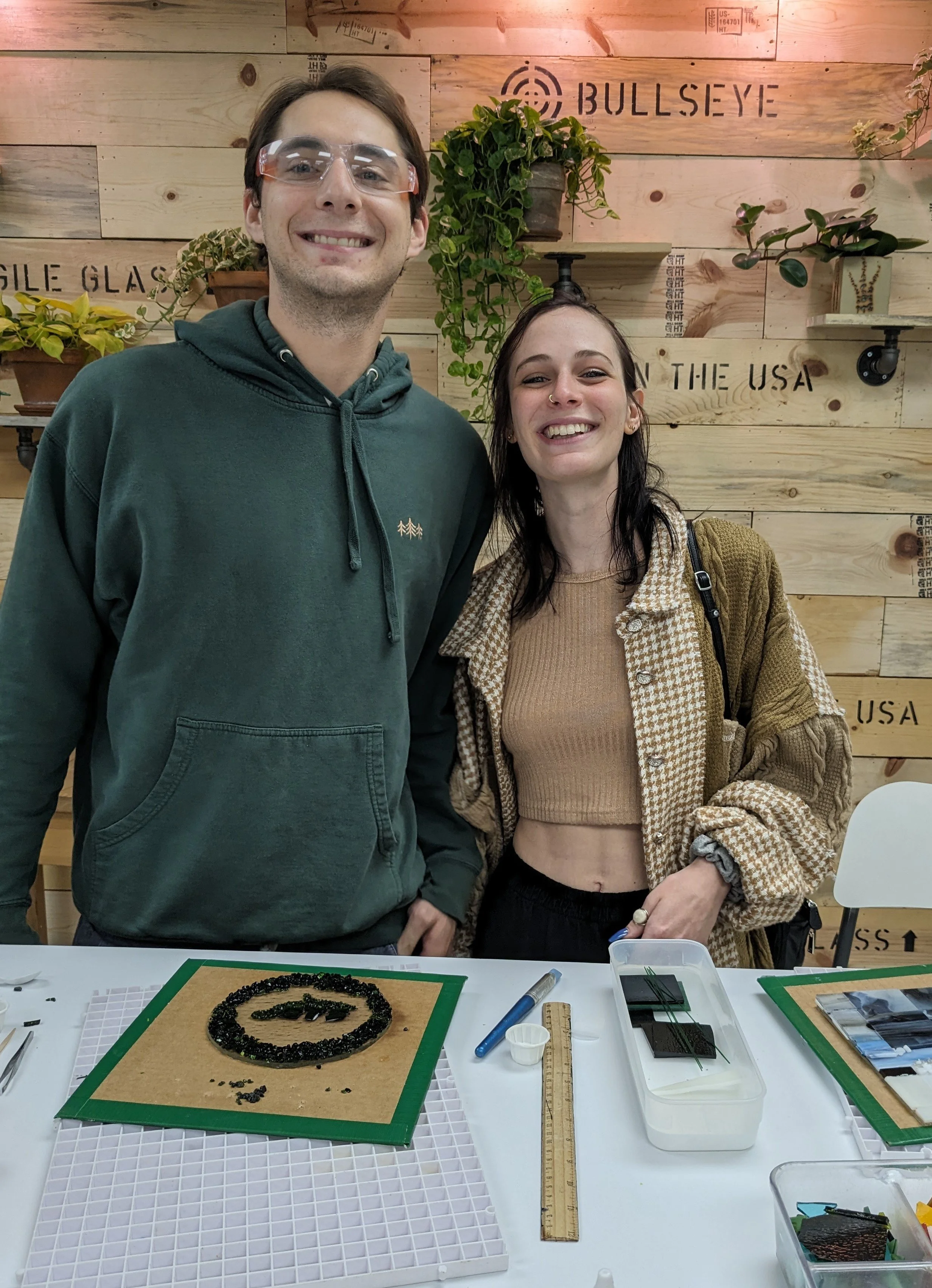 Two smiling people stand behind a table with craft materials, with a wooden wall background decorated with plants and signs related to the USA.