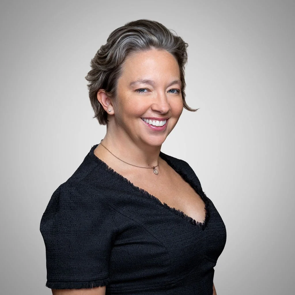 A woman with short, gray hair and a bright smile, wearing a black top and a delicate necklace, standing against a plain gray background.