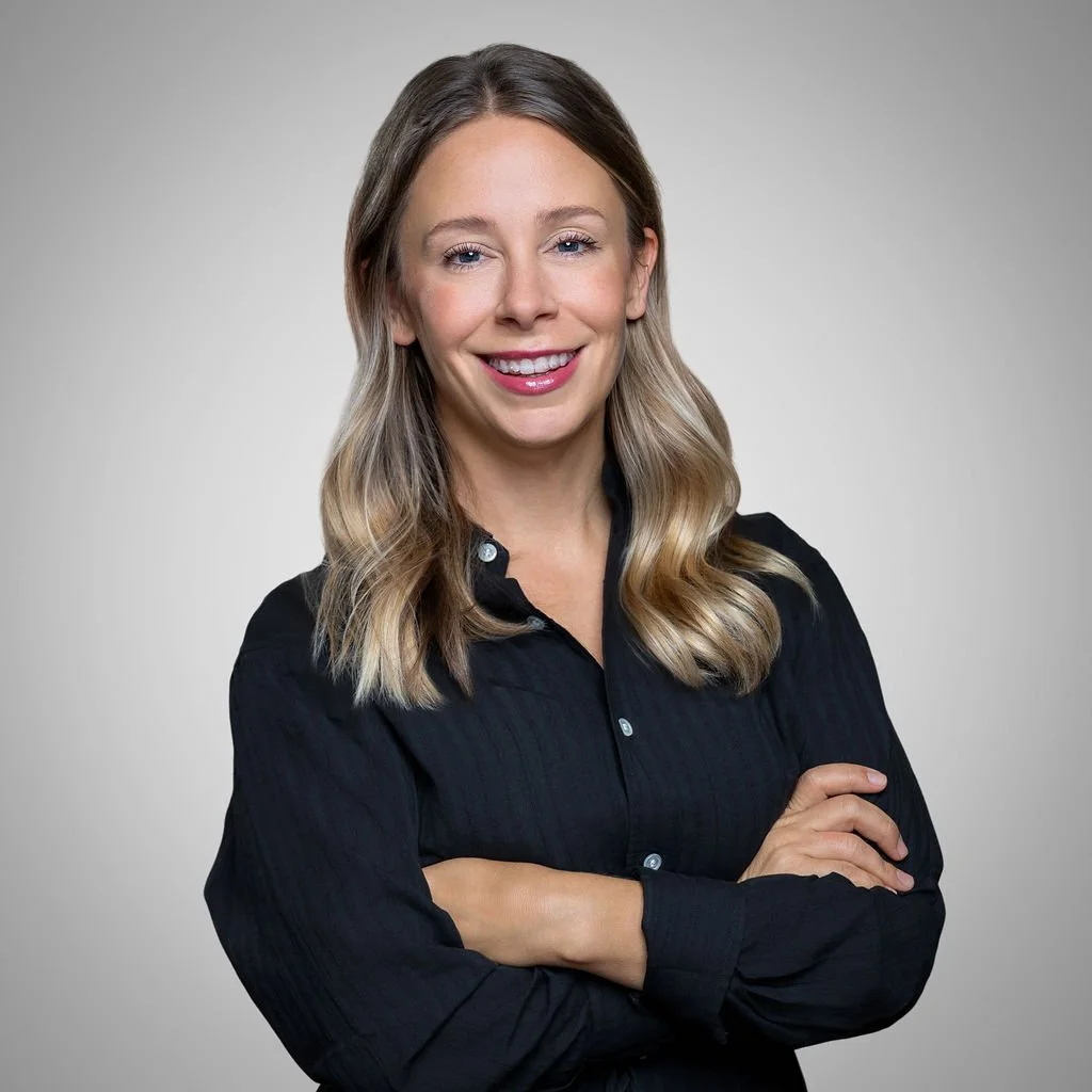 A woman with blonde wavy hair wearing a black shirt, smiling with arms crossed against a plain gray background.