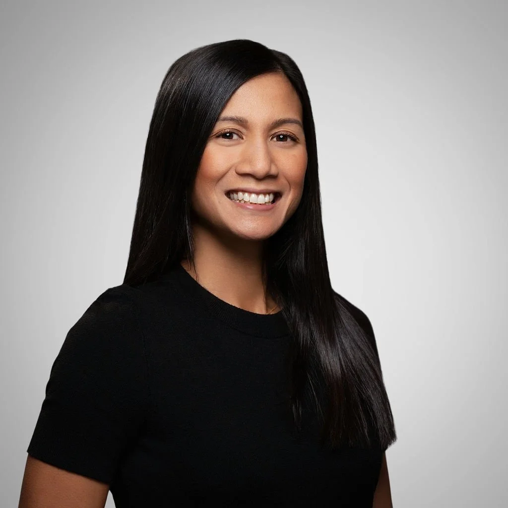 A young woman with long black hair smiling, wearing a black shirt, against a plain gray background.