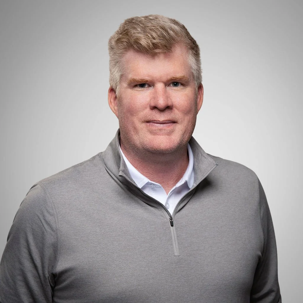 Portrait of a man with blond hair wearing a gray zip-up jacket over a white collared shirt, smiling against a plain light gray background.