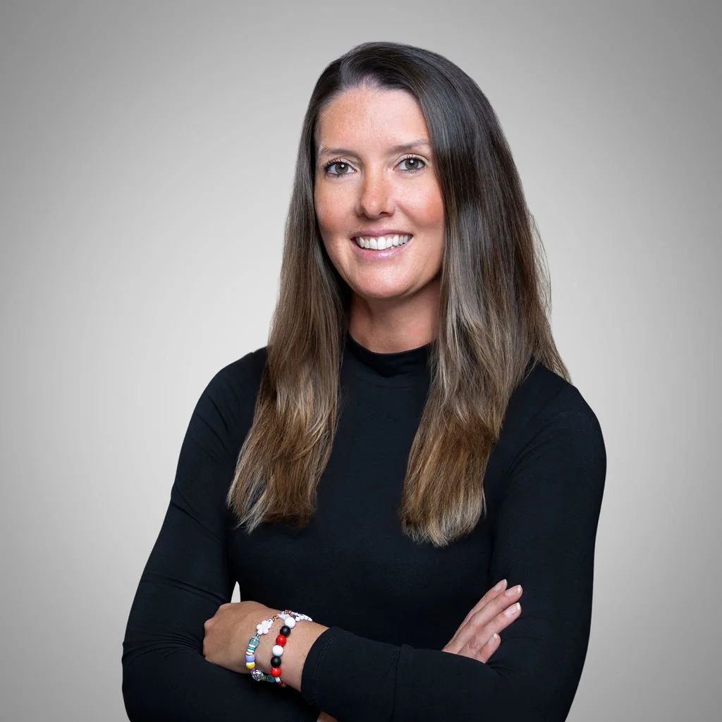 A woman with long brown hair, wearing a black long-sleeve top, smiling, with her arms crossed, standing against a plain light gray background.