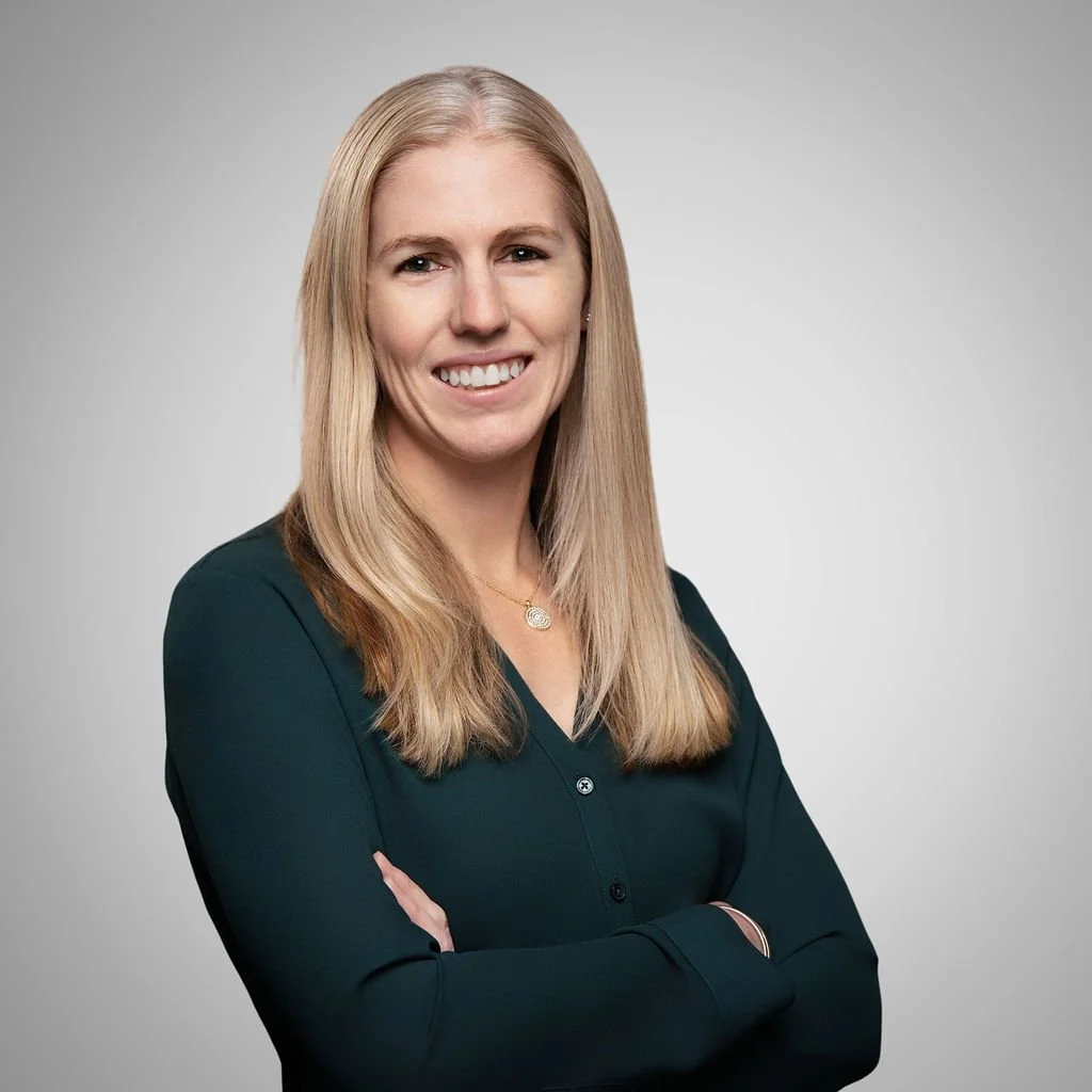 A woman with long blonde hair smiling, wearing a dark green blouse, and standing against a light gray background.
