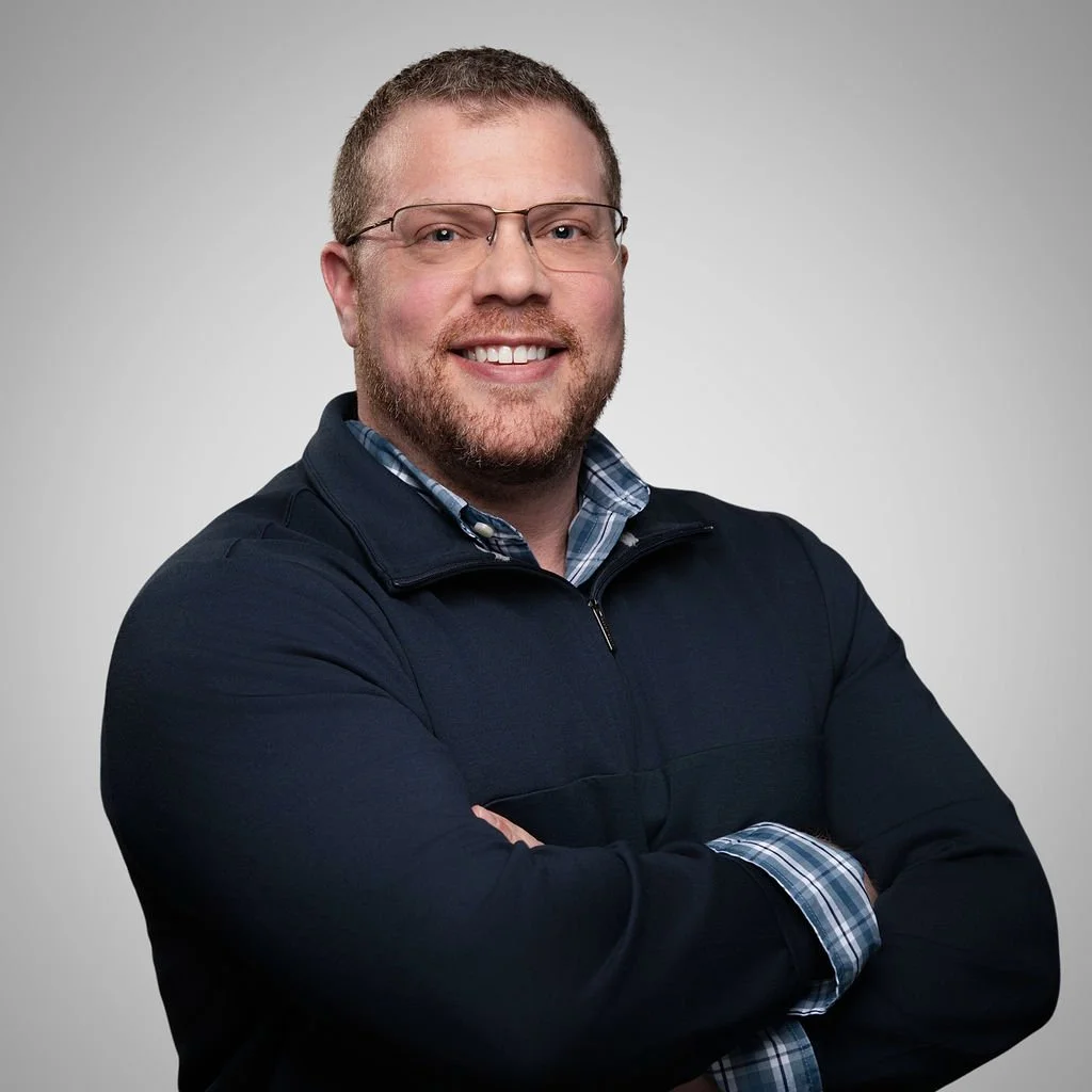 A man with glasses and a beard, smiling with arms crossed, wearing a dark jacket and a checkered shirt, against a plain gray background.