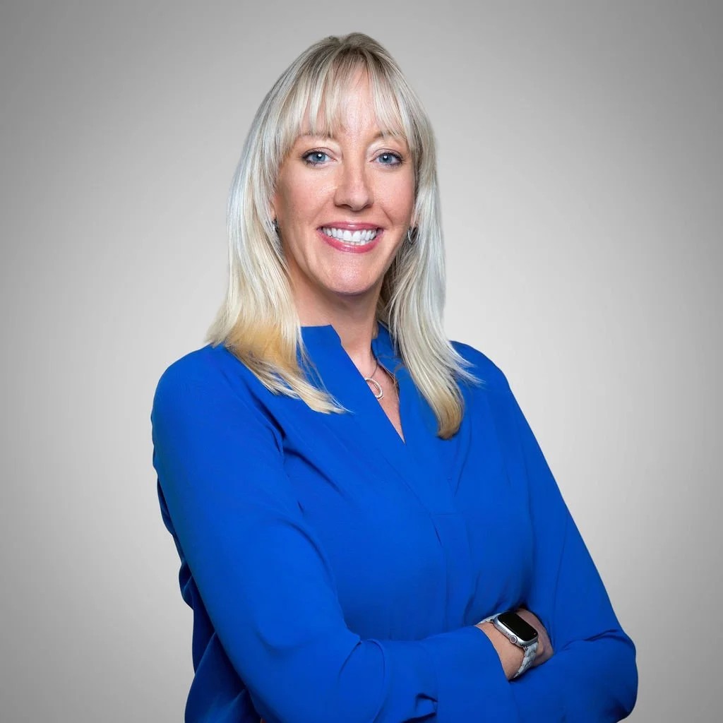 A woman with blonde hair and blue eyes wearing a bright blue blouse, smiling, with arms crossed, standing against a plain grey background.